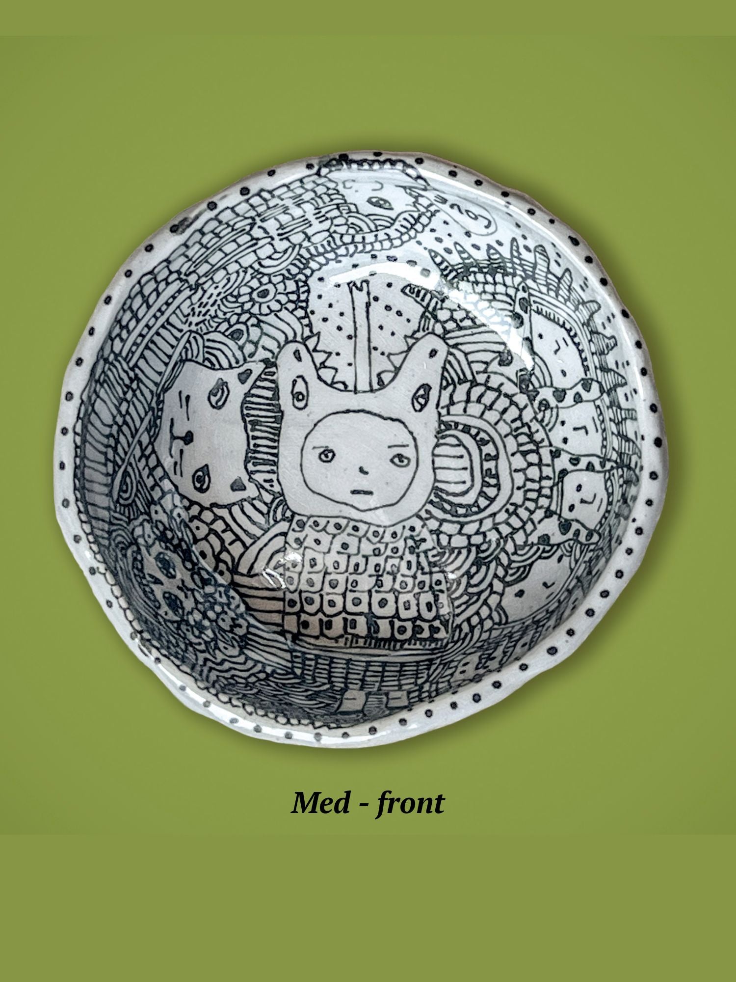 Decorative bowl with black and white patterns on a green background. A face is in the center surrounded by a mix of lines and other faces. The rim is dotted.