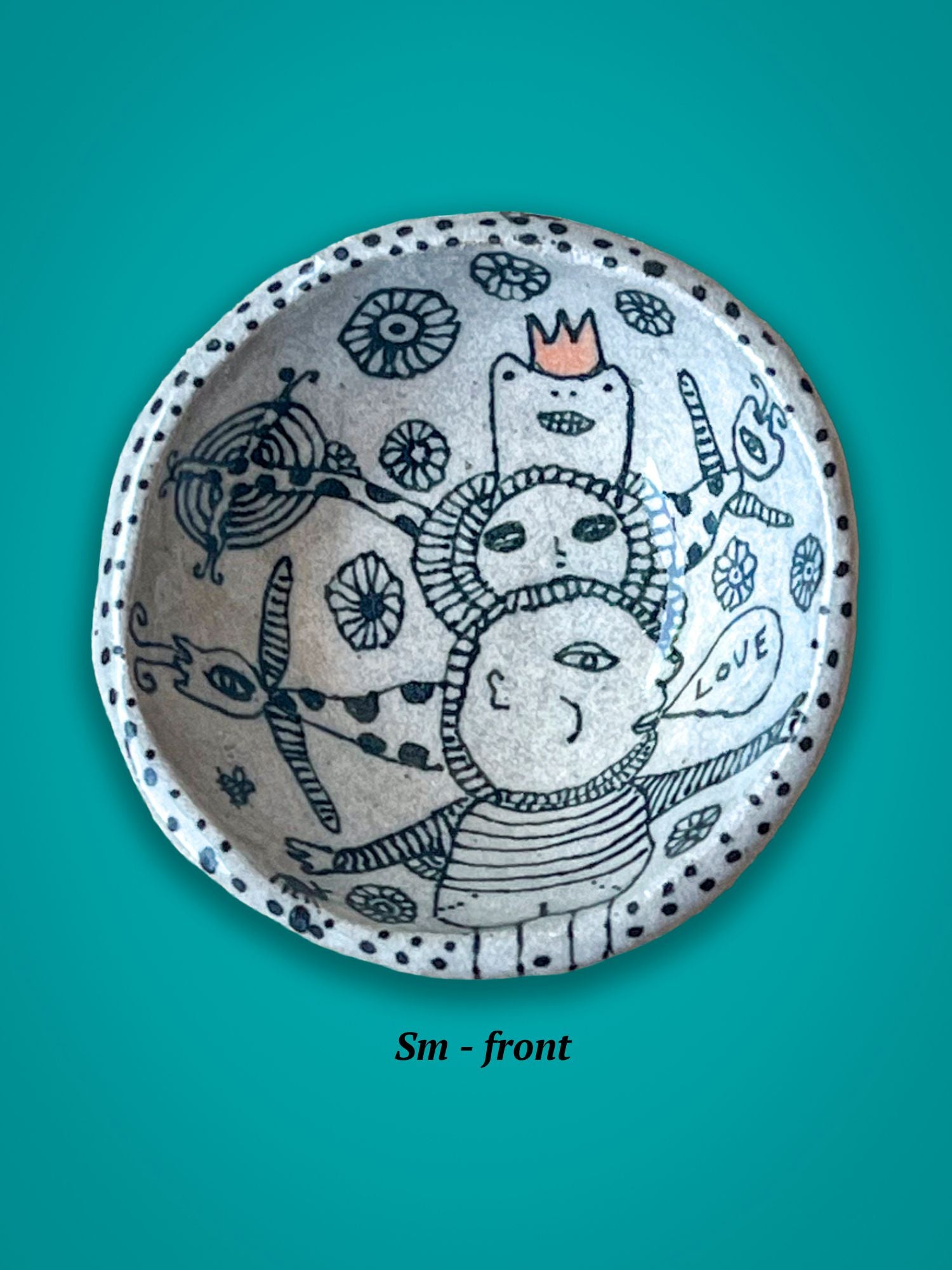 Ceramic bowl with artistic design on a teal background. There are several whimsical figures, one has an orange crown against the white and black images.