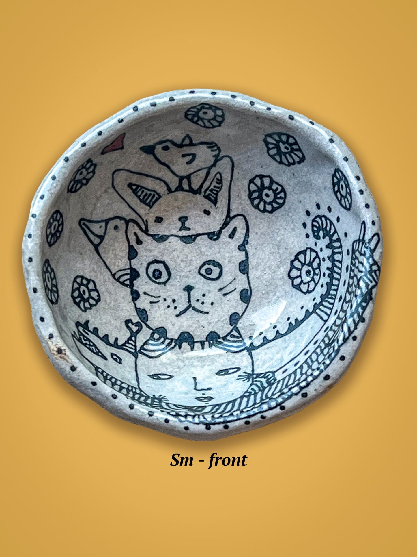 Ceramic bowl with cat illustrations rests on a yellow background. The cats are layered and a bird sits on top.