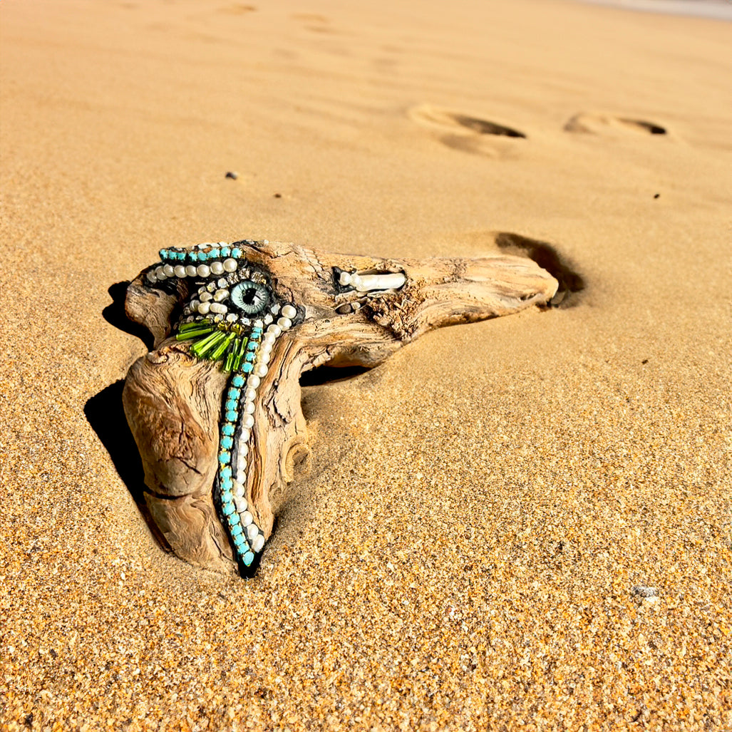 Natural driftwood sculpture decorated with beads, stones, and glass eye by Denise Marshall title 'Bird's Eye View' in a sandy setting; 6"Wx4"H