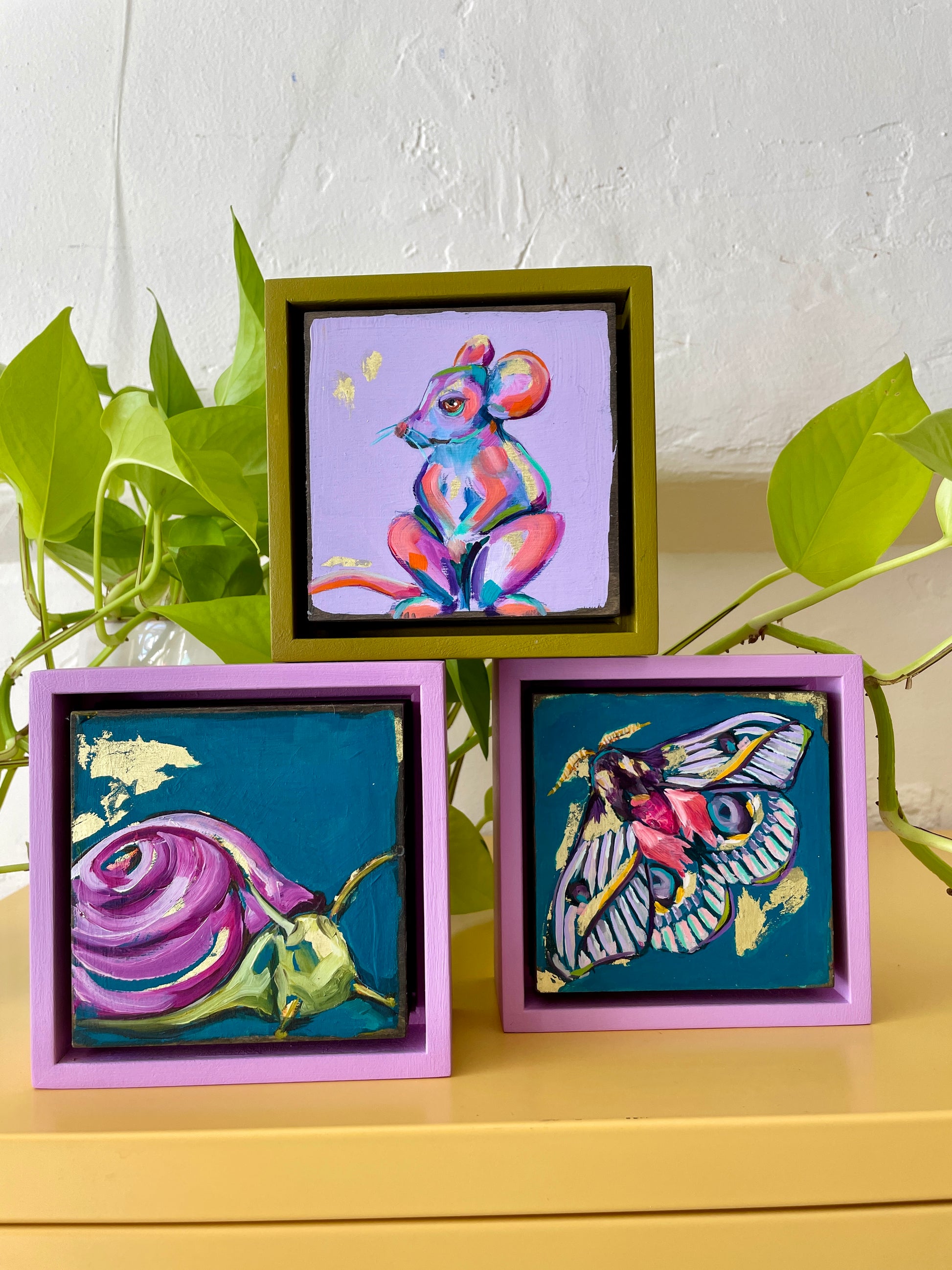 Set of three colorful paintings, a mouse, a moth, and a snail placed on a table; purples and blues with touches of gold leaf; each 5"x5" with frame; artist Shaney Watters