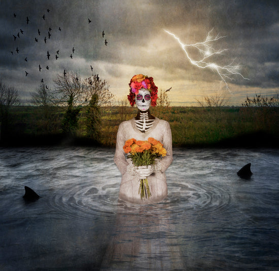 A Day of the Dead Catrina rises from a pond  holding a bouquet of flowers. Two shark fins can be seen in the water as a storm and lightening fill the sky.