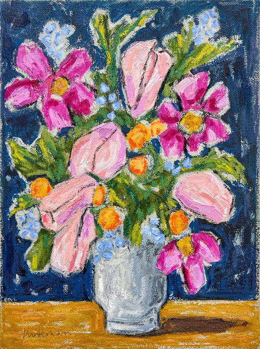 This is a beautiful still life of a mixture of flowers in a white base against a blue background. The flowers are of a mixed variety in pinks, oranges and dark pink. We also see the green foliage.