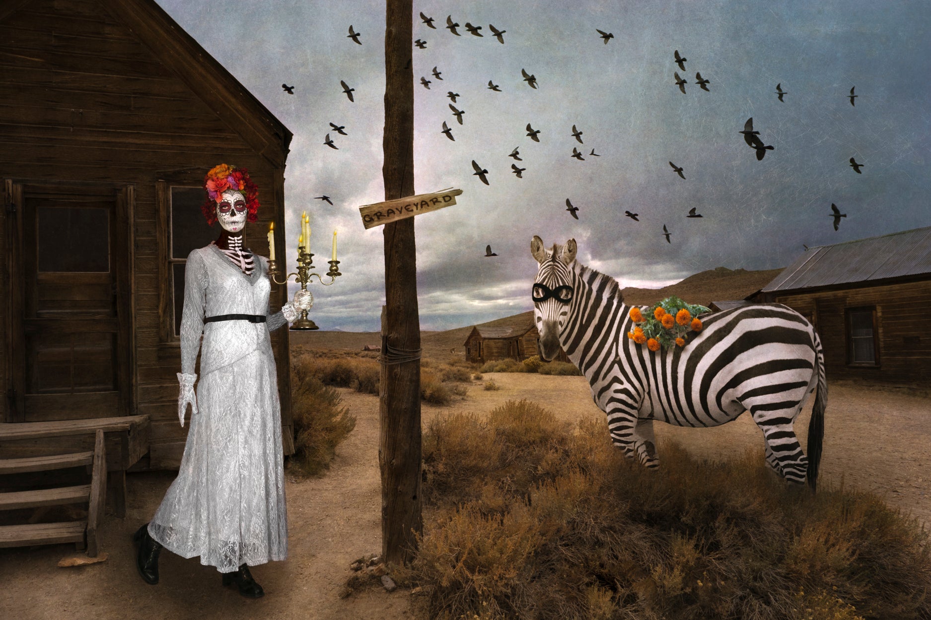 A Catrina skeleton is dressed all in white with a red headress. She is holding a candelabra and contemplating  a zebra with a bouquet of flowers on its back. The setting is a deserted ghost town and a murder of crows flies above in a stormy sky.