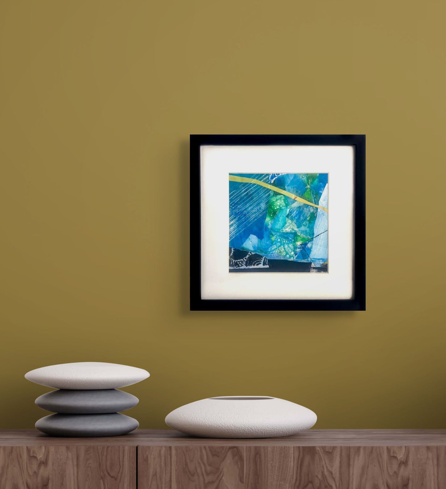 Framed 8"x8" abstract artwork with blue, green, and yellow colors; white mat, 1" black frame; artist Bob Hogue; shown on light brown wall in situ