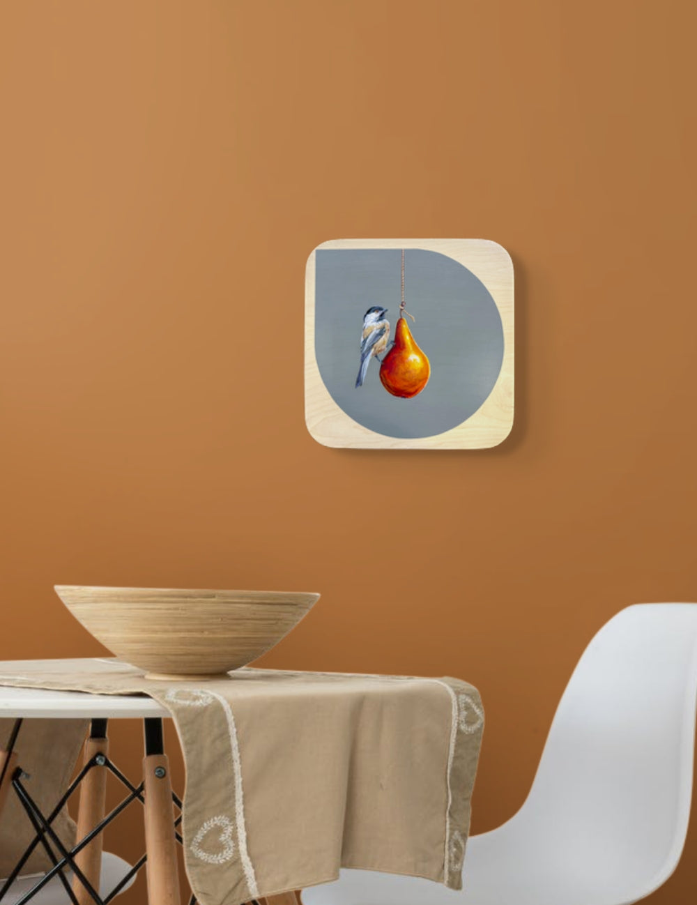 Dark blue, tan, and white Blackcapped Chickadee perched on pear hanging from light string; gray background on natural wood panel; shown in situ on orange wall above table and chair; Marie Lavallee artist