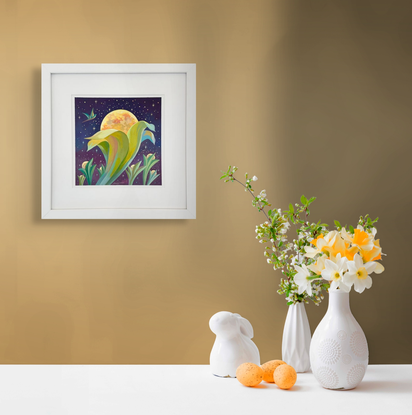 This in-situ image shows a framed Blooming Moons hanging over a white table. On the table sit two small flower vases, three orange eggs and a small white porcelain rabbit