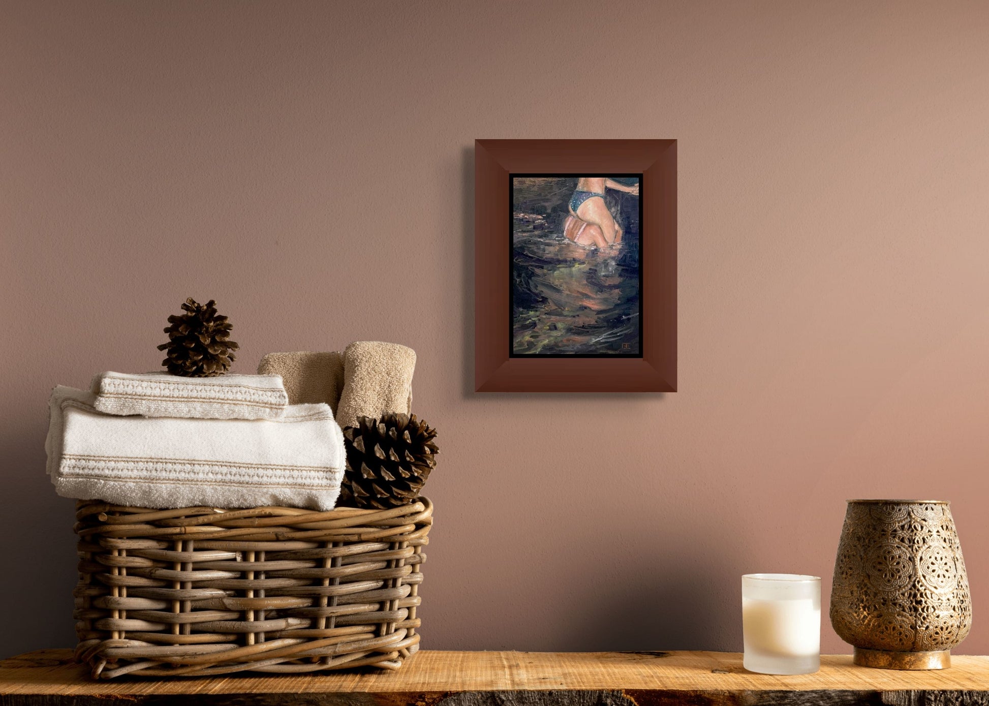 Framed here in a medium brown frame with an inner black border for an in-situ image, the painting hangs on a taupe wall over a table with a basket of towels and pine cones. A candle and vase sit to the right side on the table.