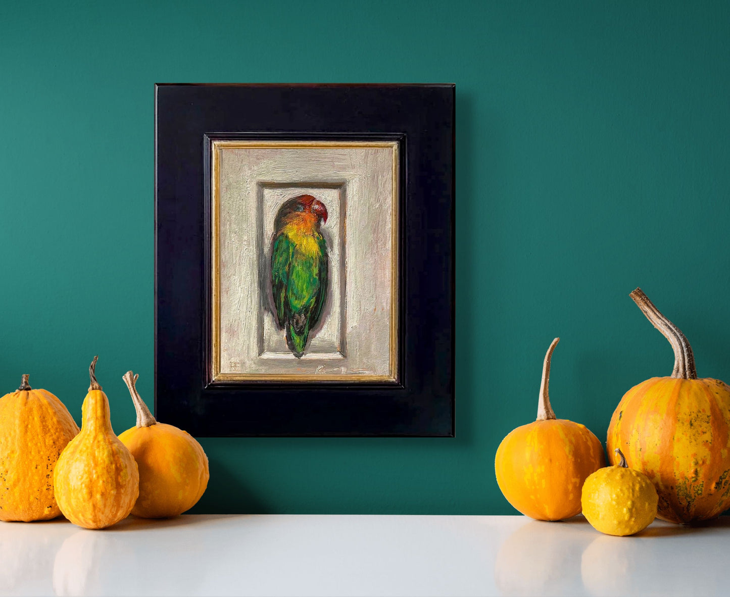 Painting of a colorful dead parrot lying in a small enclosure on a textured light brown background; shown with 2" black wood frame; in situ hanging on teal wall above yellow squash on countertop.