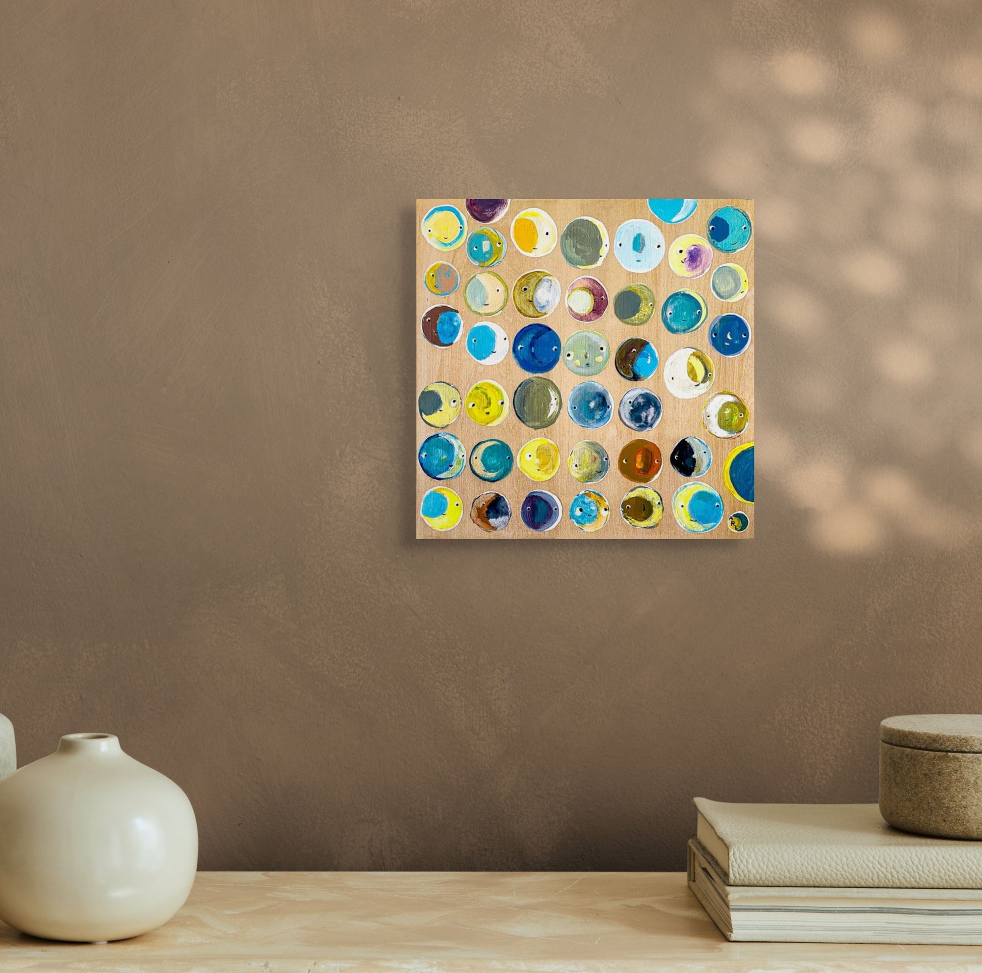 A painting featuring a number of small moons of many colors and faces. The moons are arranged in a random pattern in tones of blues, yellows, teals and greens; shown in situ on brown wall