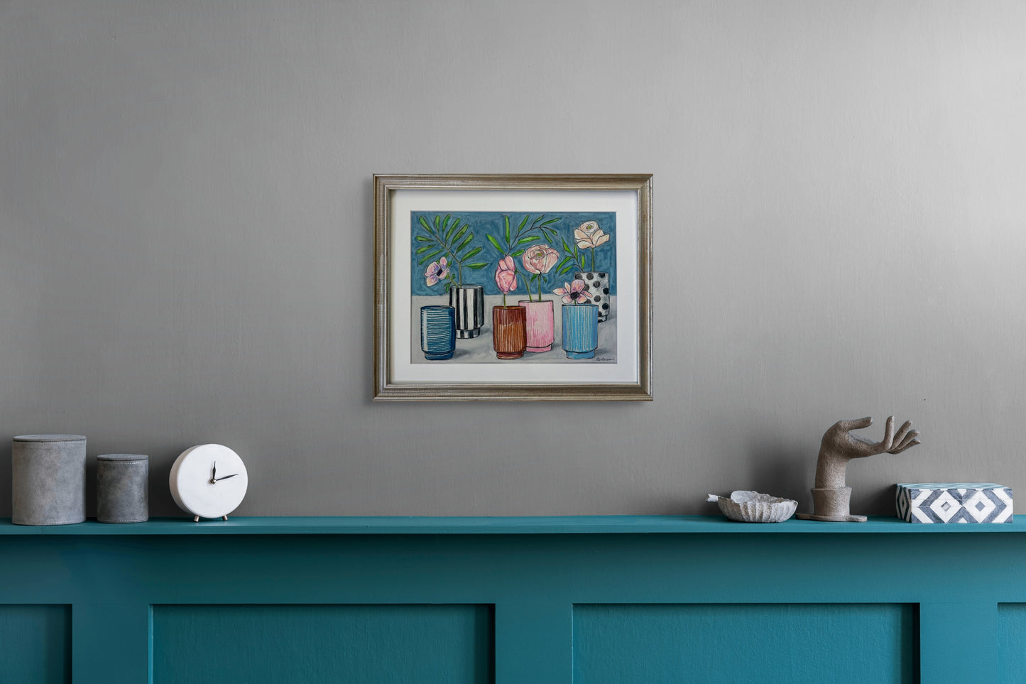 Fill Your Cups is shown hanging on a light gray wall over a teal blue counter with several items including a clock and a sculpture of a hand.