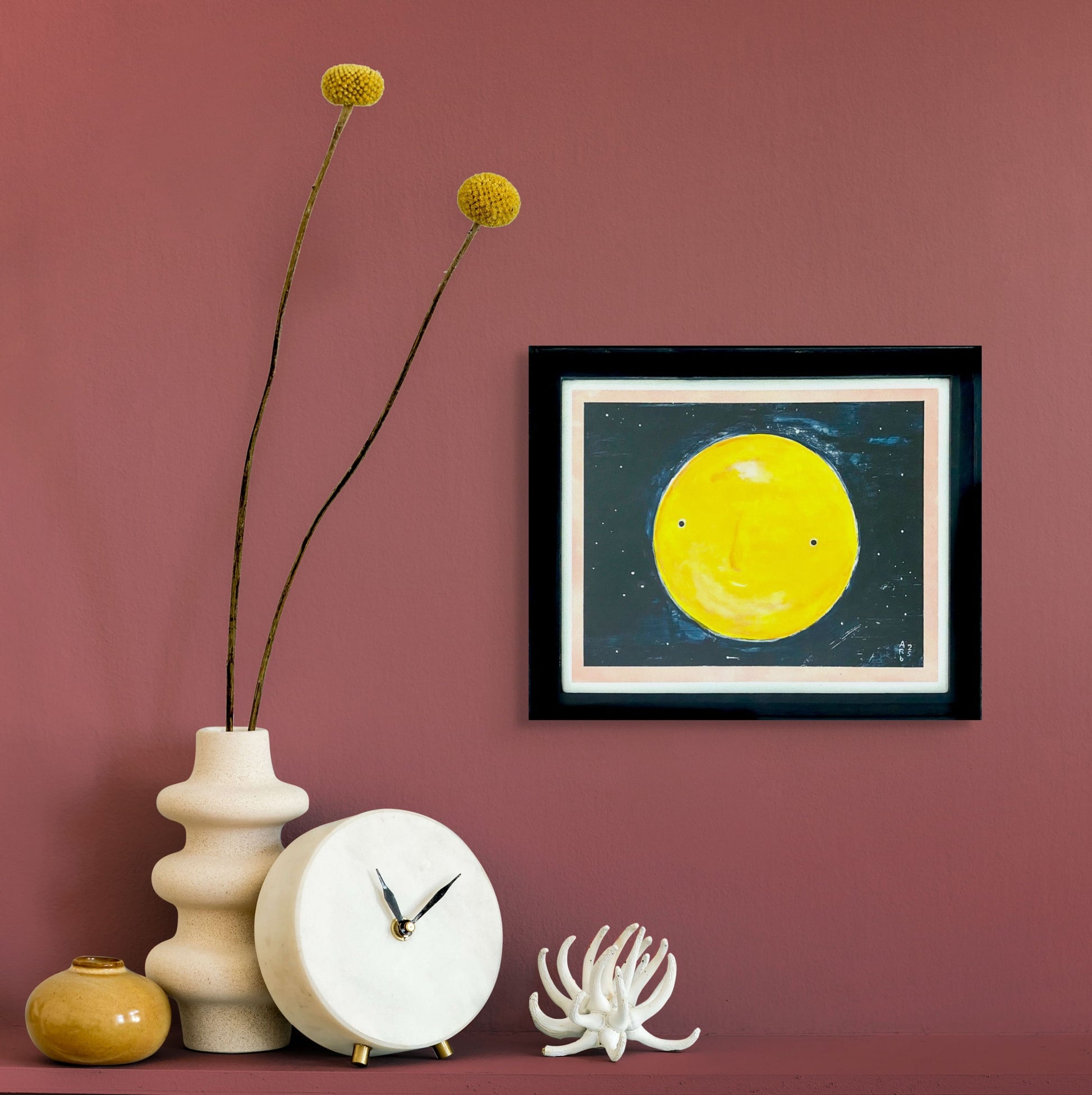 This full yellow moon has a face with a sly smile. It is set on the background of a starry night sky; it is framed  in black wood with a white & pink border around the image; shown in situ against red wall and shelf w/clock & vases
