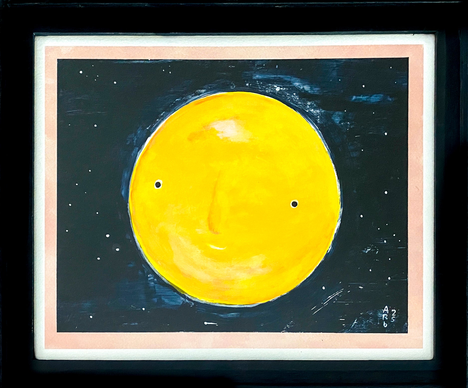 This full yellow moon has a face with a sly smile. It is set on the background of a starry night sky; it is framed  in black wood with a white & pink border around the image.