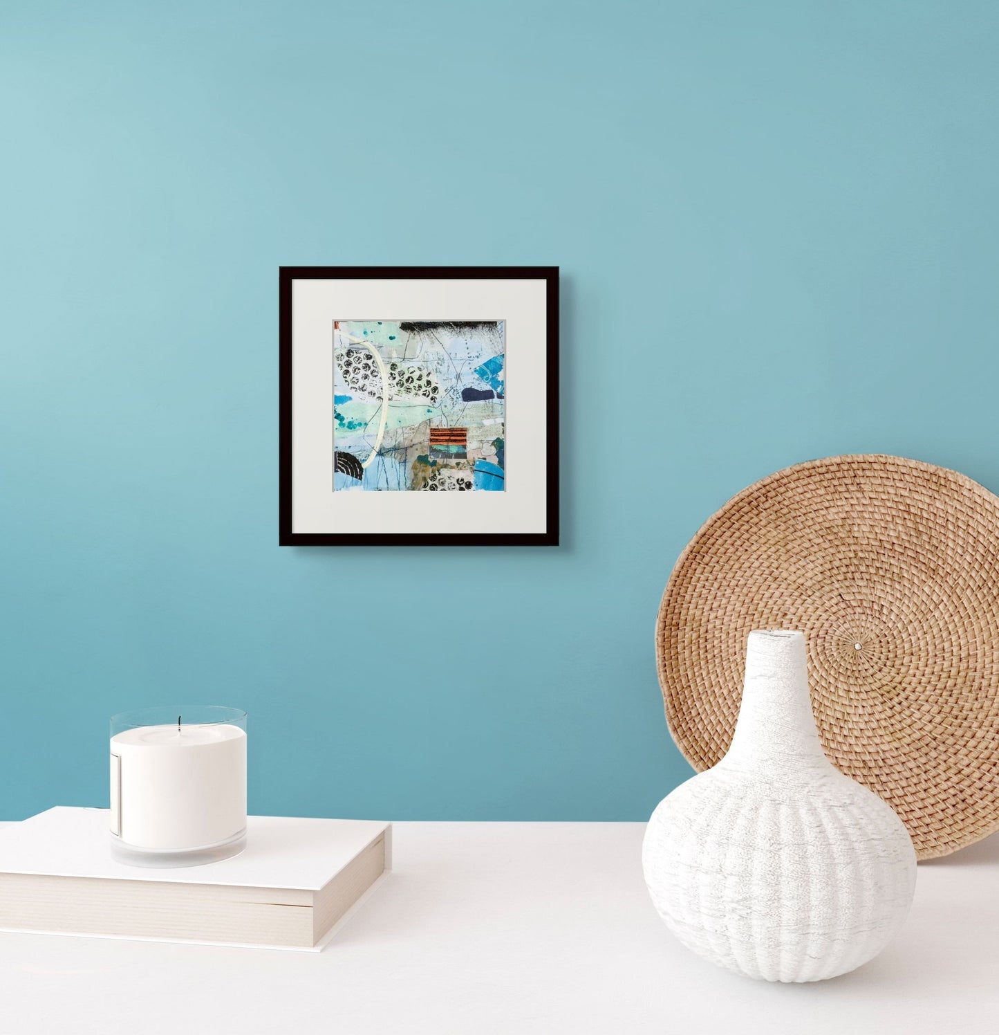 Lasting Impressions is shown here framed and matted and hanging on a light teal wall over a table with a vase, a candle and basket.