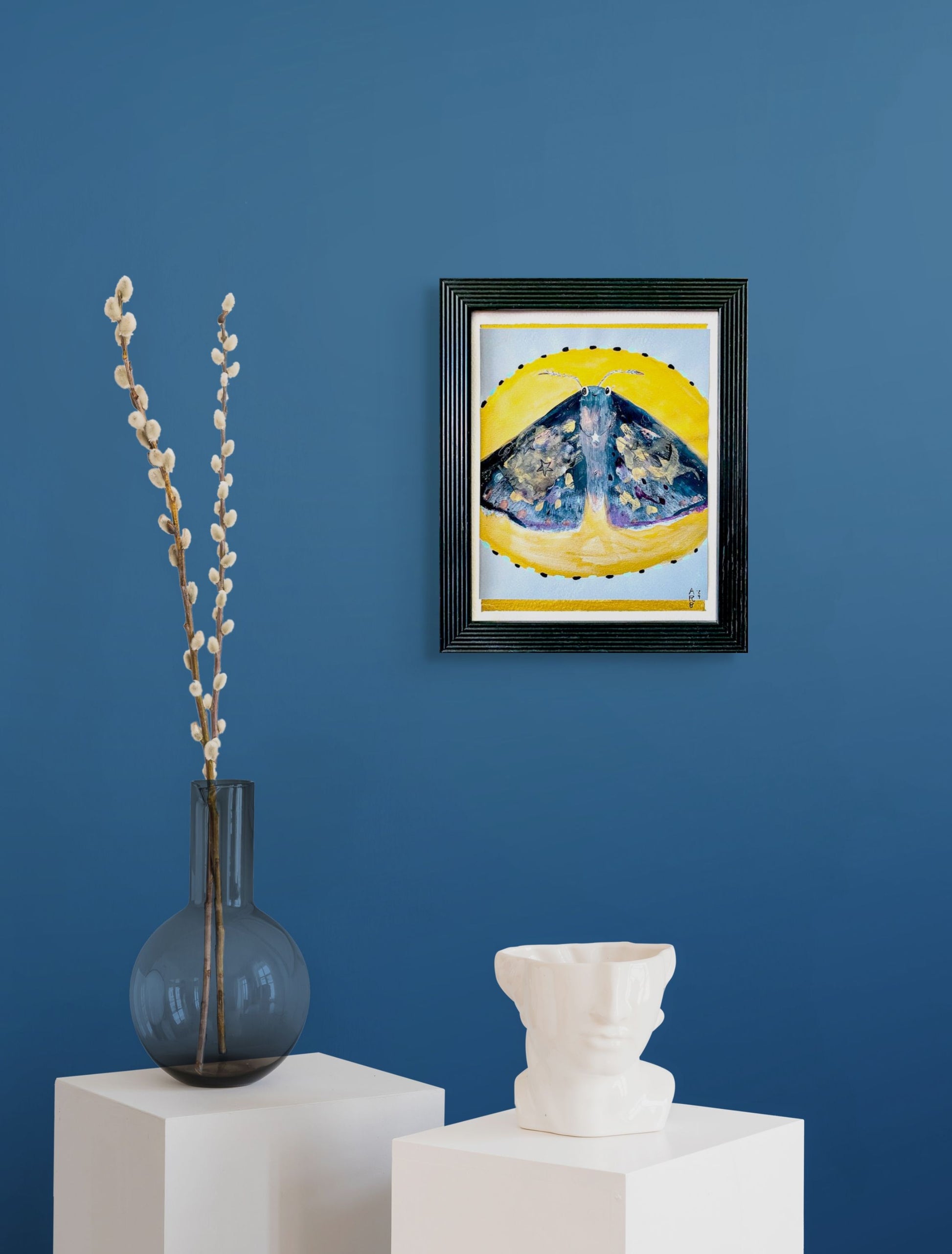 Framed artwork of a beautiful moth against the backdrop of a yellow moon on a light blue background; shown in situ against blue wall w/several sculptures