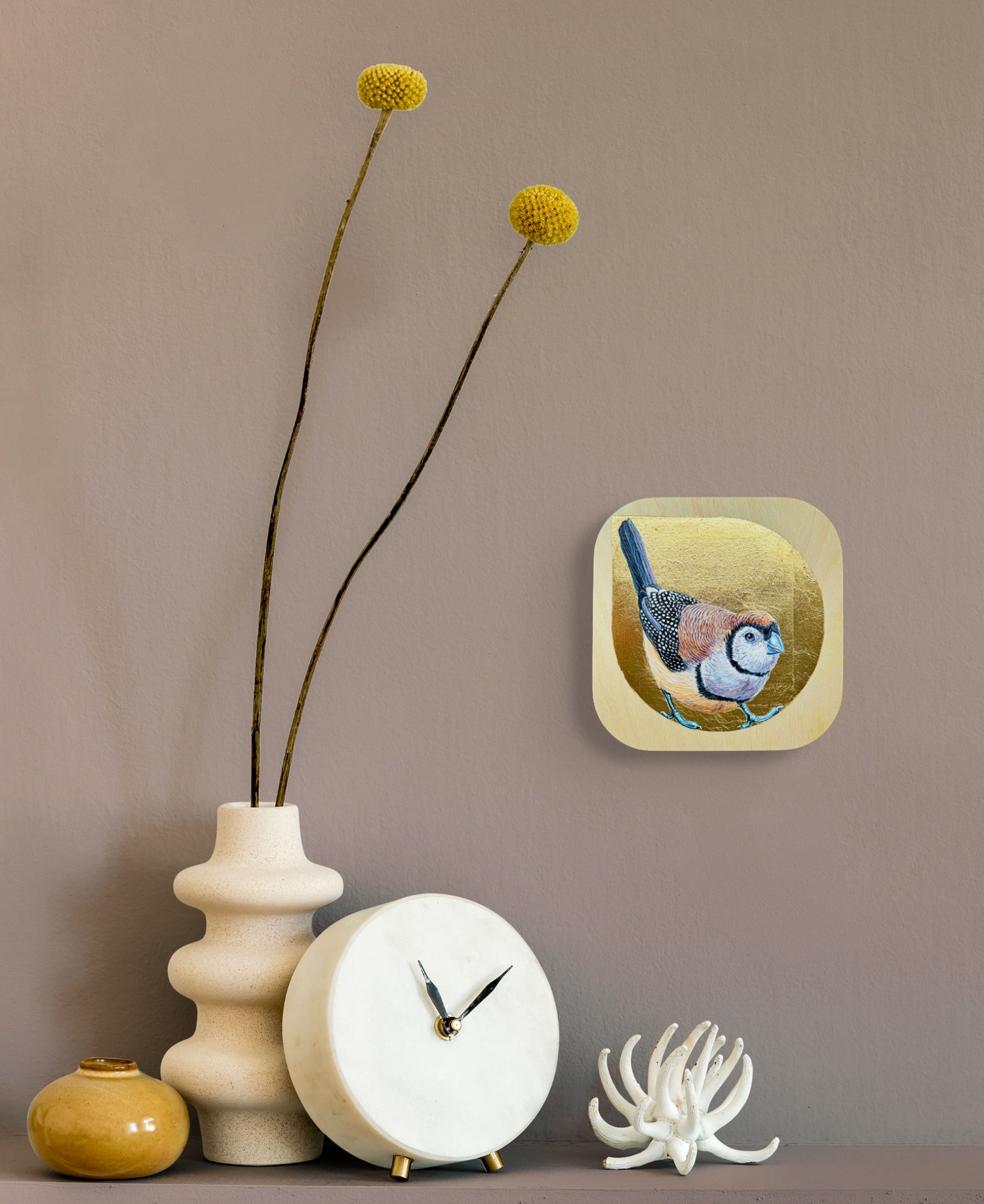 The painting hangs on a light taupe wall over a collection of items on a shelf including a small wooden spool vase, a clock and a shell.