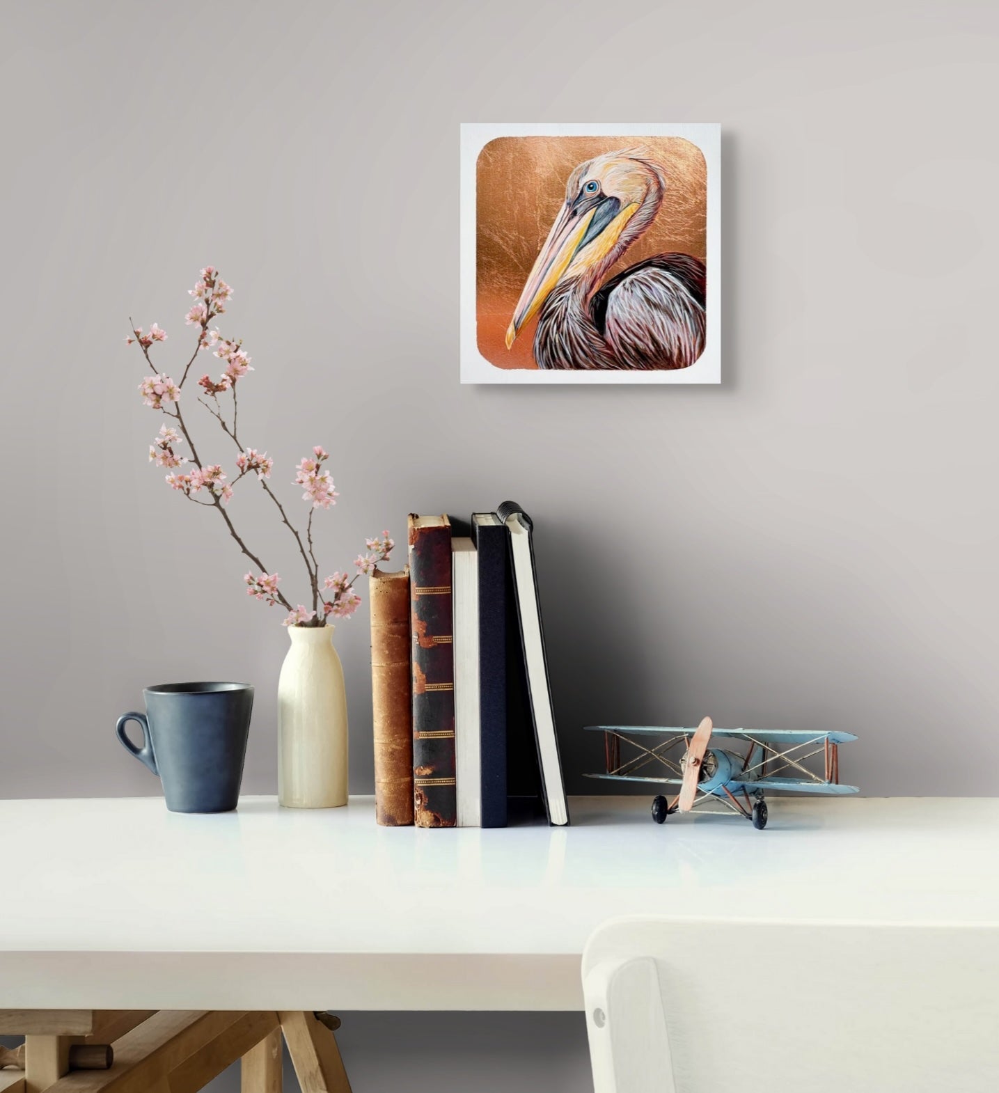 A copper leaf background works  with the hues of the feathering on this image of a pelican's face and beak. The layers of color which form the head and the striking blue eye makes for a stunning piece; artist Marie Lavallee; 8"x8"; shown in situ