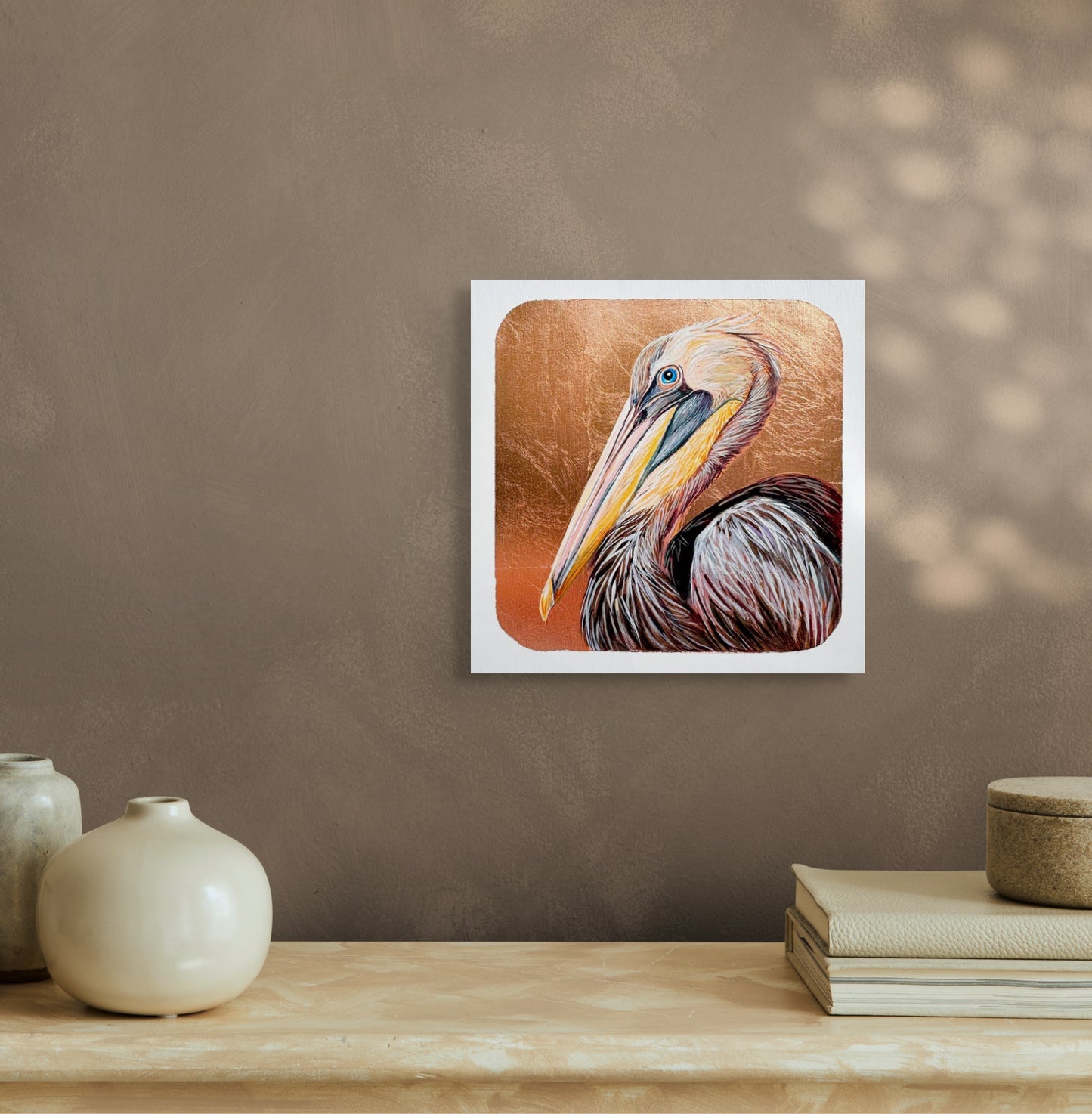 A copper leaf background works  with the hues of the feathering on this image of a pelican's face and beak. The layers of color which form the head and the striking blue eye makes for a stunning piece; artist Marie Lavallee; 8"x8"; shown in situ