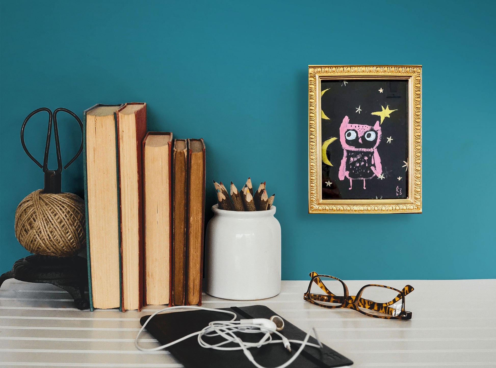 Framed artwork of Pink Owl & Two Moons shown sitting on a tabletop. The frame is gold and ornate; in situ on wall w/books and eyeglasses
