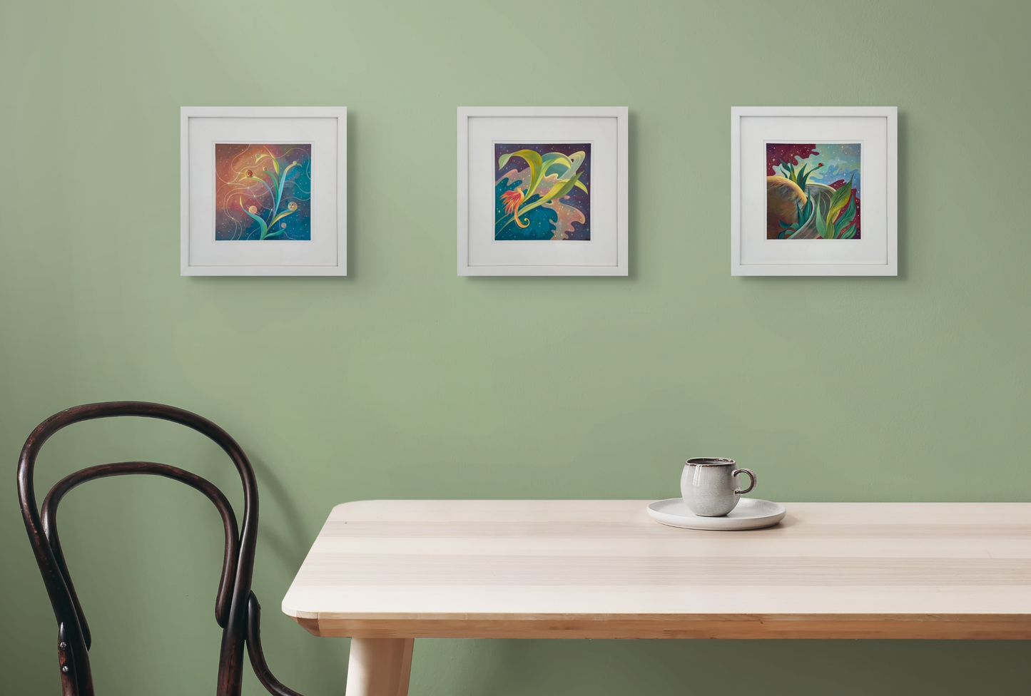 Three of Dany's framed pieces, Planetary Phases, Saturn Feathers and Lady Rings hang as a set on a light green wall above a light oak table. A mug sits on the table and a black chair is to the left.