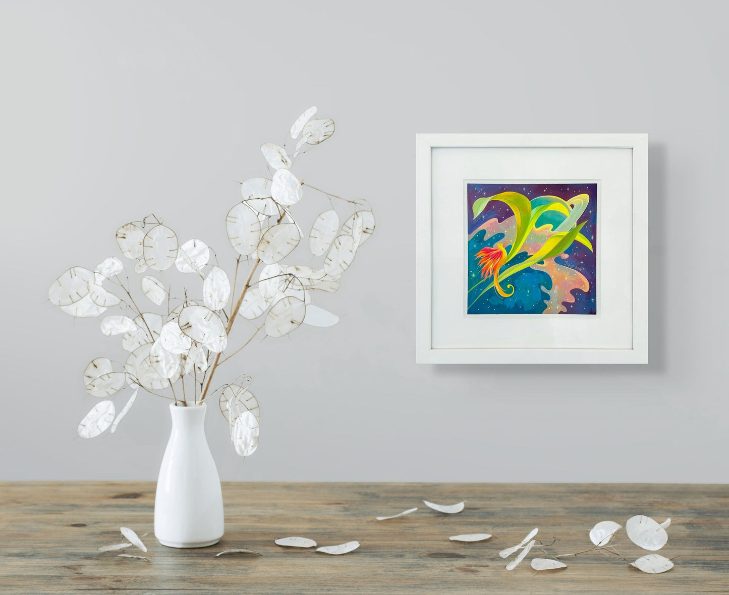 Saturn Feathers is shown here in situ on a pale grey wall. It hangs above a table adorned with a ceramic vase holding white dried leaves