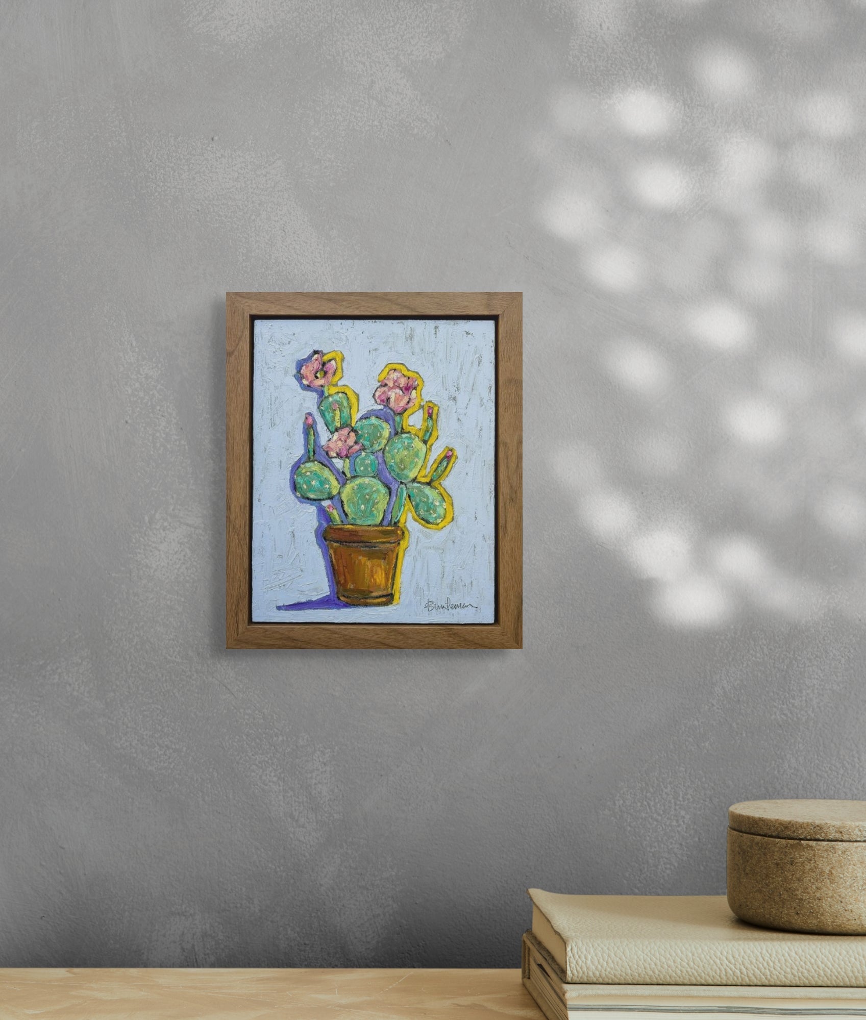 This image is in situ of the piece. It is hanging on a light gray wall and the frame is a medium shade of brown wood. Sunlight comes in from a window on the right. There is a table with a set of books on the right edge of the in situ picture.