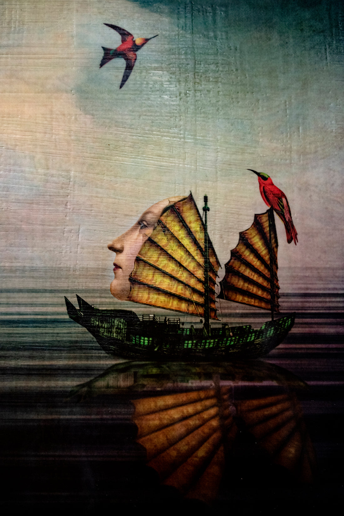A surreal composite image of a boat with a woman's face looking up at a bird in the sky.  Image is titles A Fair Wind by Meryl Skyler and has an encaustic wax finish.
