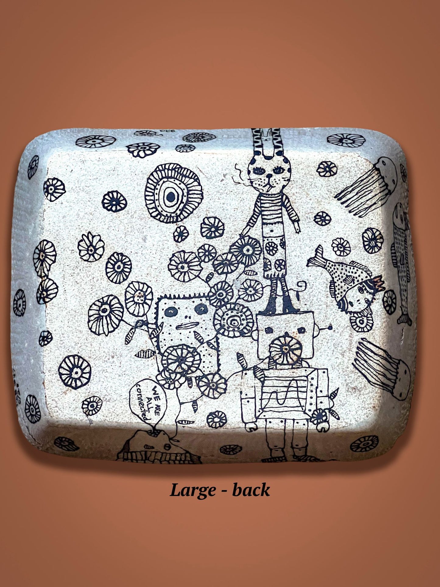 This image shows the back of the rectangular ceramic plate with black and white illustrations resting on a brown background. The drawings are of whimsical figures. This underside view is not glossy like the other side.