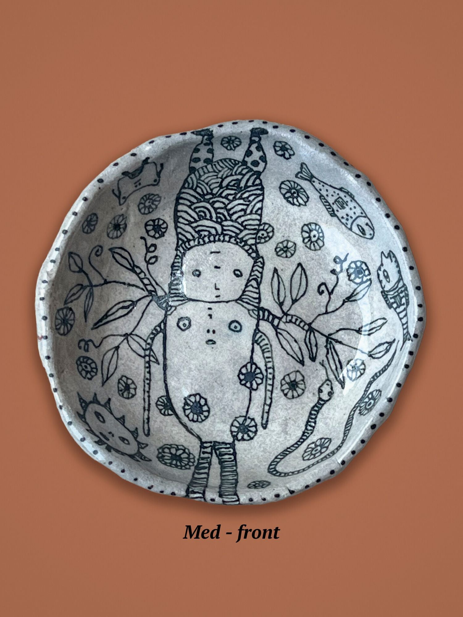 Decorative ceramic plate with black line art shown on a brown background. The figures are whimsical and look like they are connected at the top. Fish, leaves and branches fill out the design.