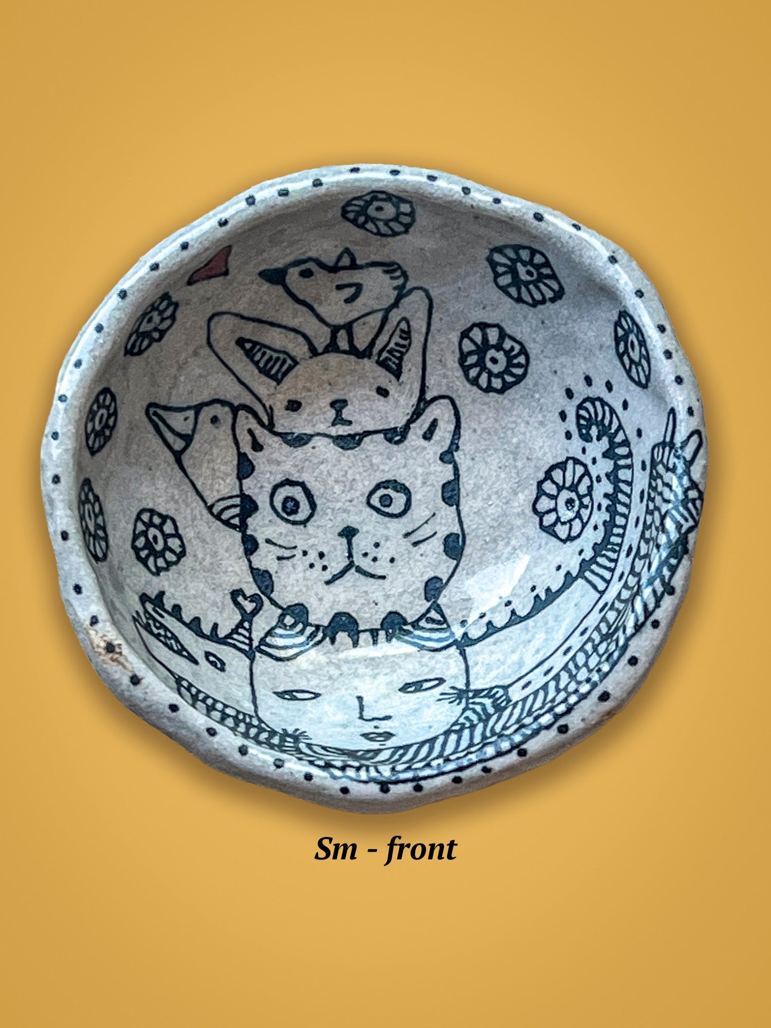 Ceramic bowl with cat illustrations rests on a yellow background. The cats are layered and a bird sits on top. 
