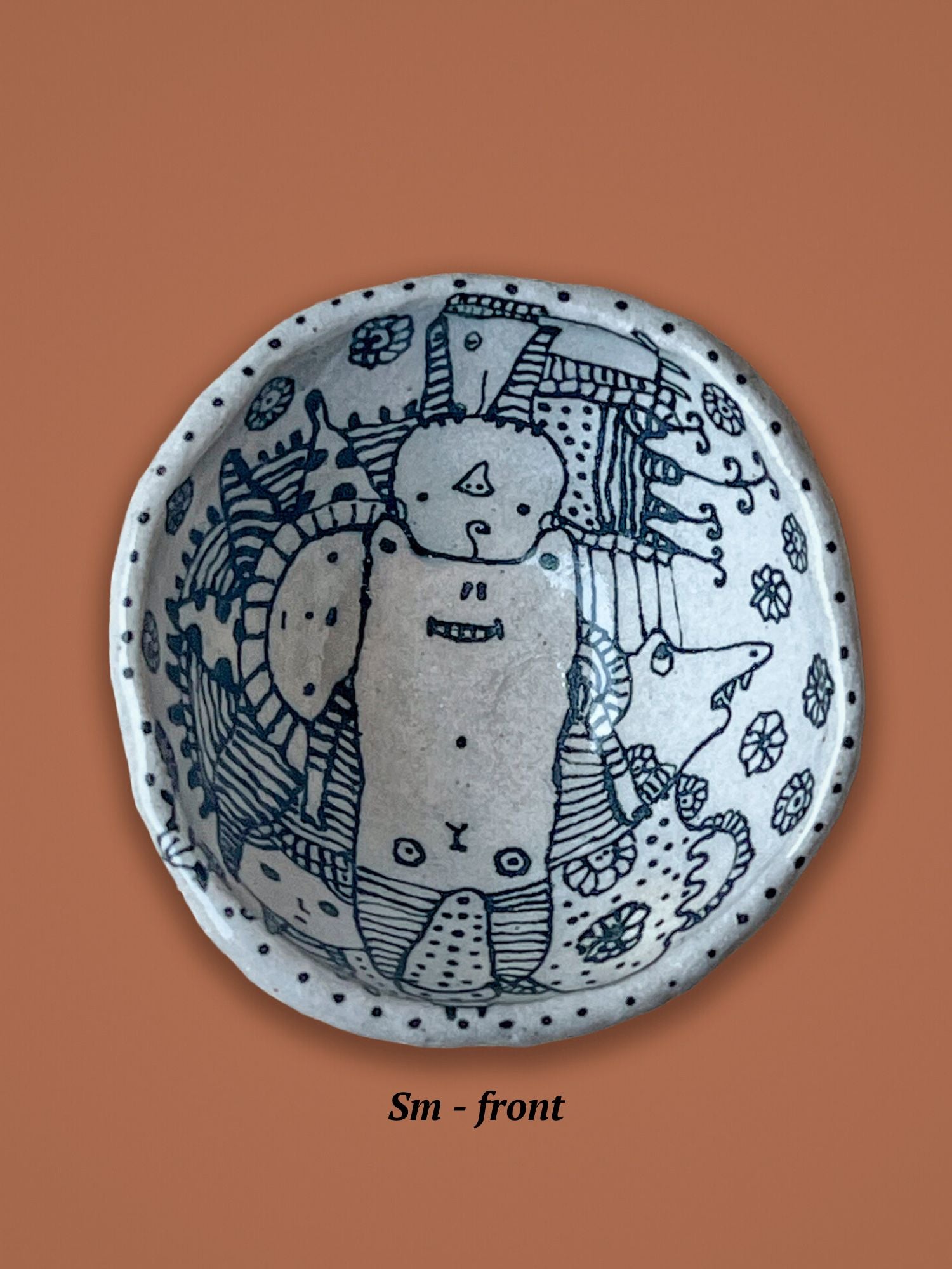 Ceramic plate with black line art shown on a brown background. The bowl has whimsical characters hand drawn and surrounded by hand drawn flowers. 