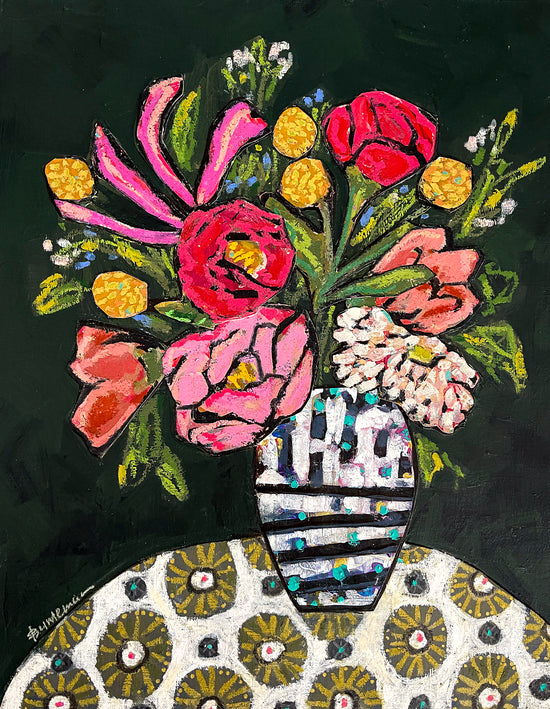 A vase of colorful flowers, mostly red, pink and yellow, are held in a white and teal patterned vase. The vase sits atop a table cloth with a circle pattern in ivory, brown and teal. 