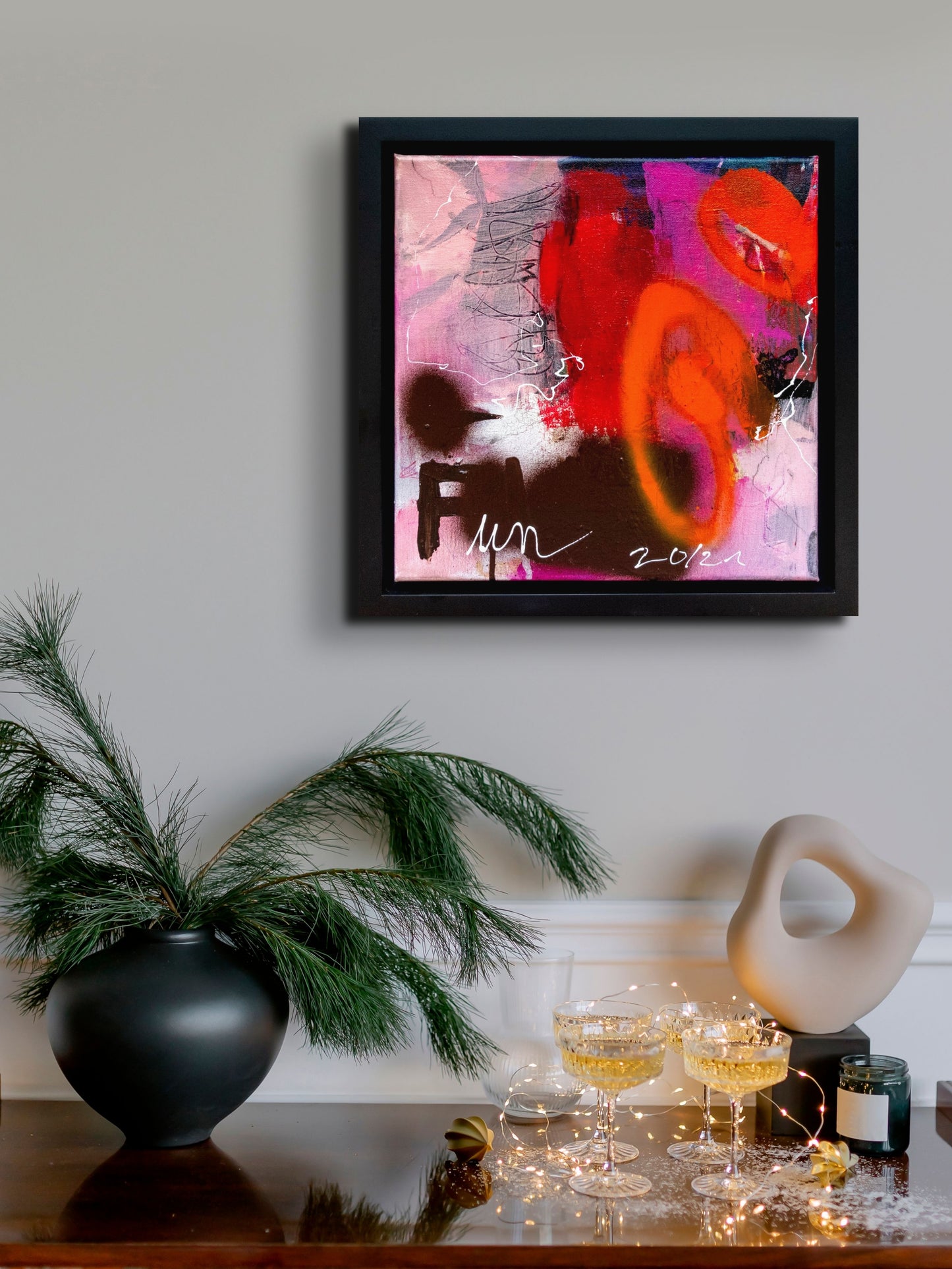 Colorful abstract painting using acrylic paint titled 'Fun' by artist Steffi Möllers; measures 12"x12" and 14"x14" with black framed; in situ on wall with champagne glasses and lights.