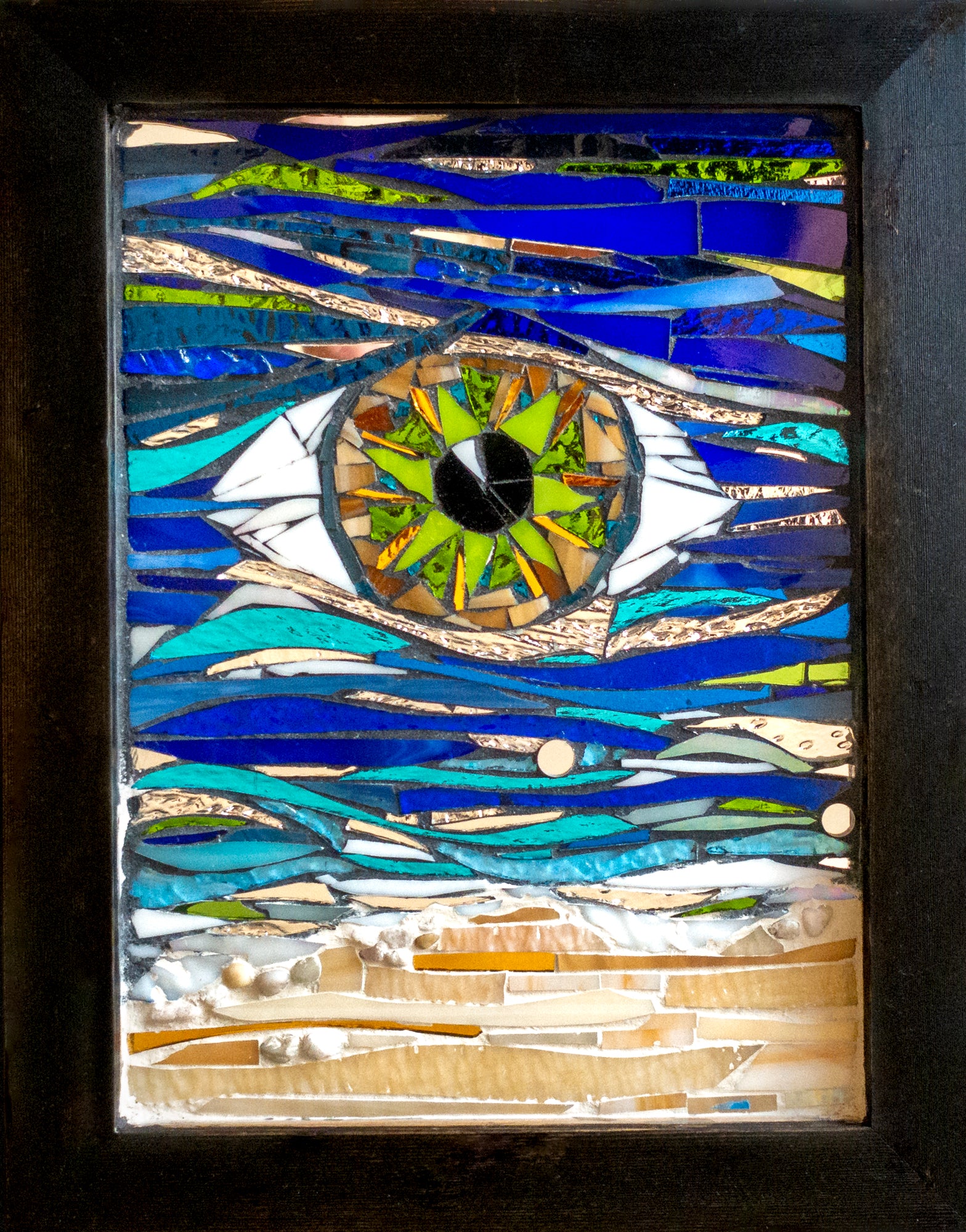 This stunning  11"x14" (with frame) mosaic piece is housed in a black frame. In the center is a green eye with touches of tans and lighter shades of green. The eye sits among a sea of blue and green glass that portray an ocean's current; artist Denise Marshall