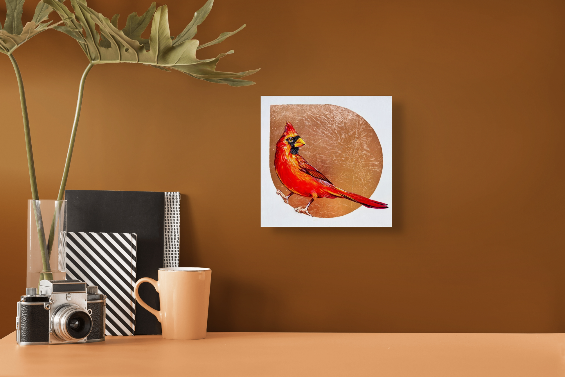 Drop of Red hangs above a small table adorned with journals, a camera, a plant and mug. The back wall is rust brown.