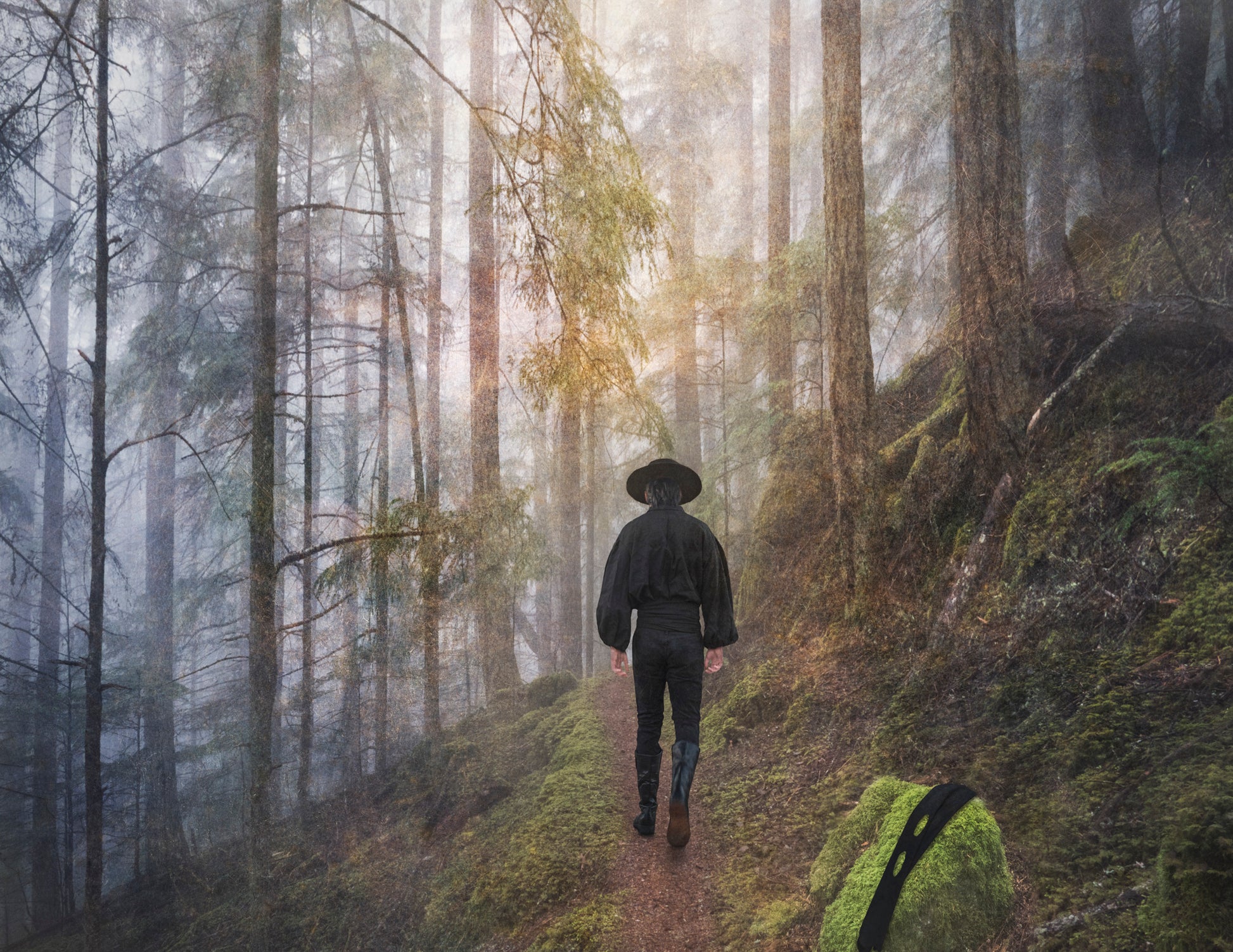  A man, clad all in black and with the signature hat worn by Zorro walks along a path through a misty forest. He has left his mask on a moss covered rock. 