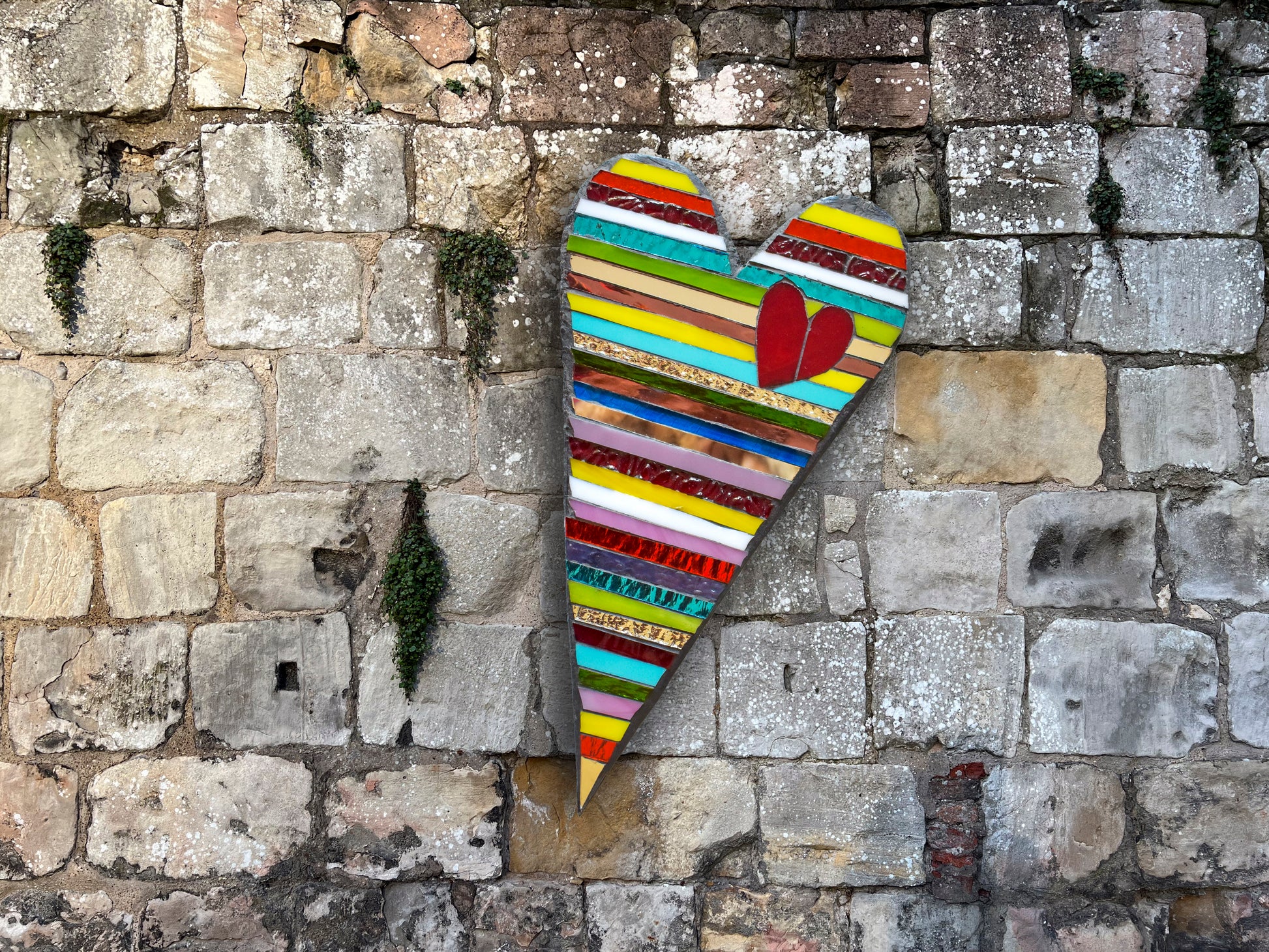 Glass mosaic heart featuring various colors of the rainbow with vertical stripes and a small red heart on the top; Artist Denise Marshall; 7.5"Wx13.5"H; shown in situ