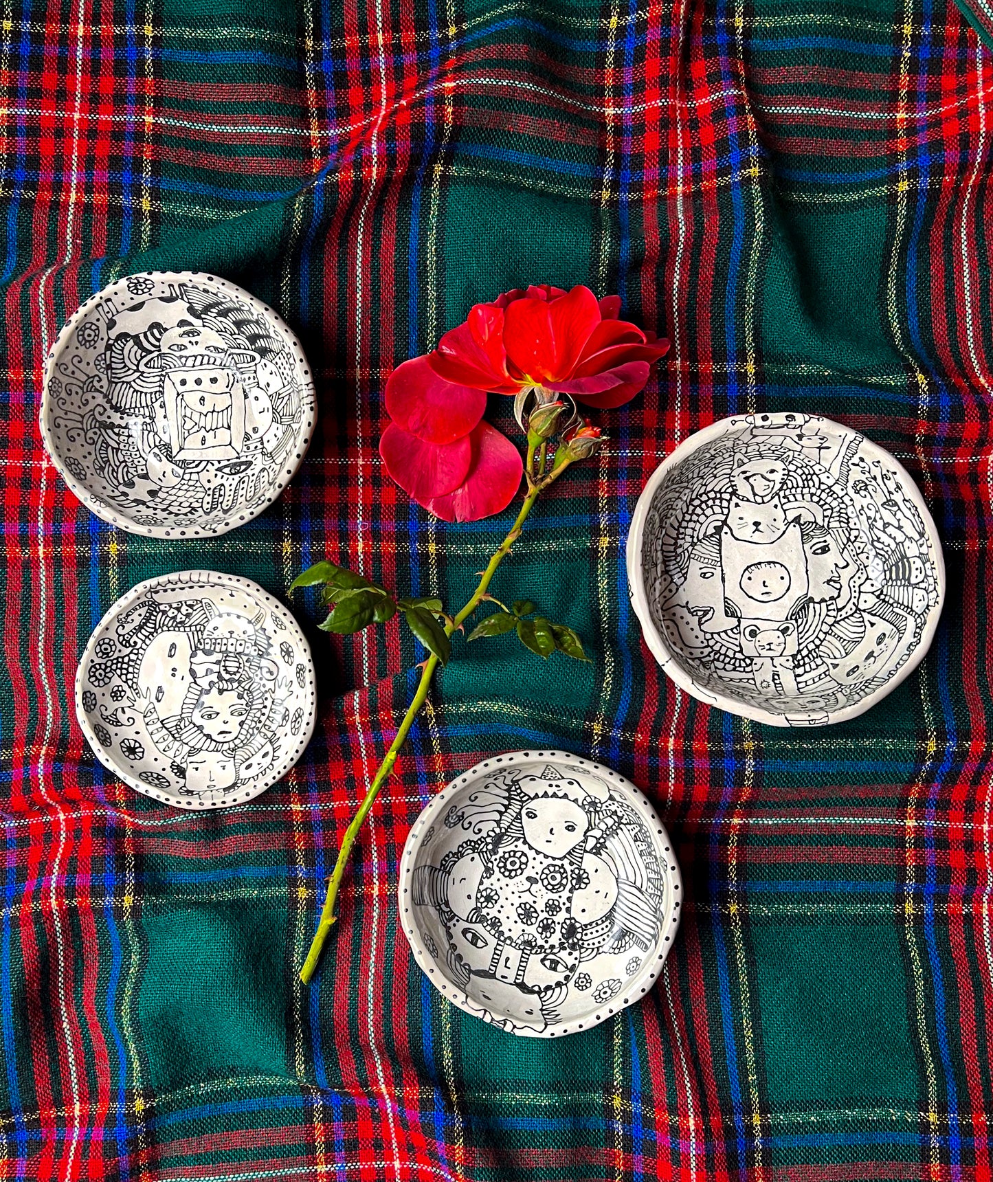 Four ceramic bowls shown together on plaid background with flower for scale with eclectic ink-drawn designs of animals, flowers, geometrics; artist Deb Blanco;  