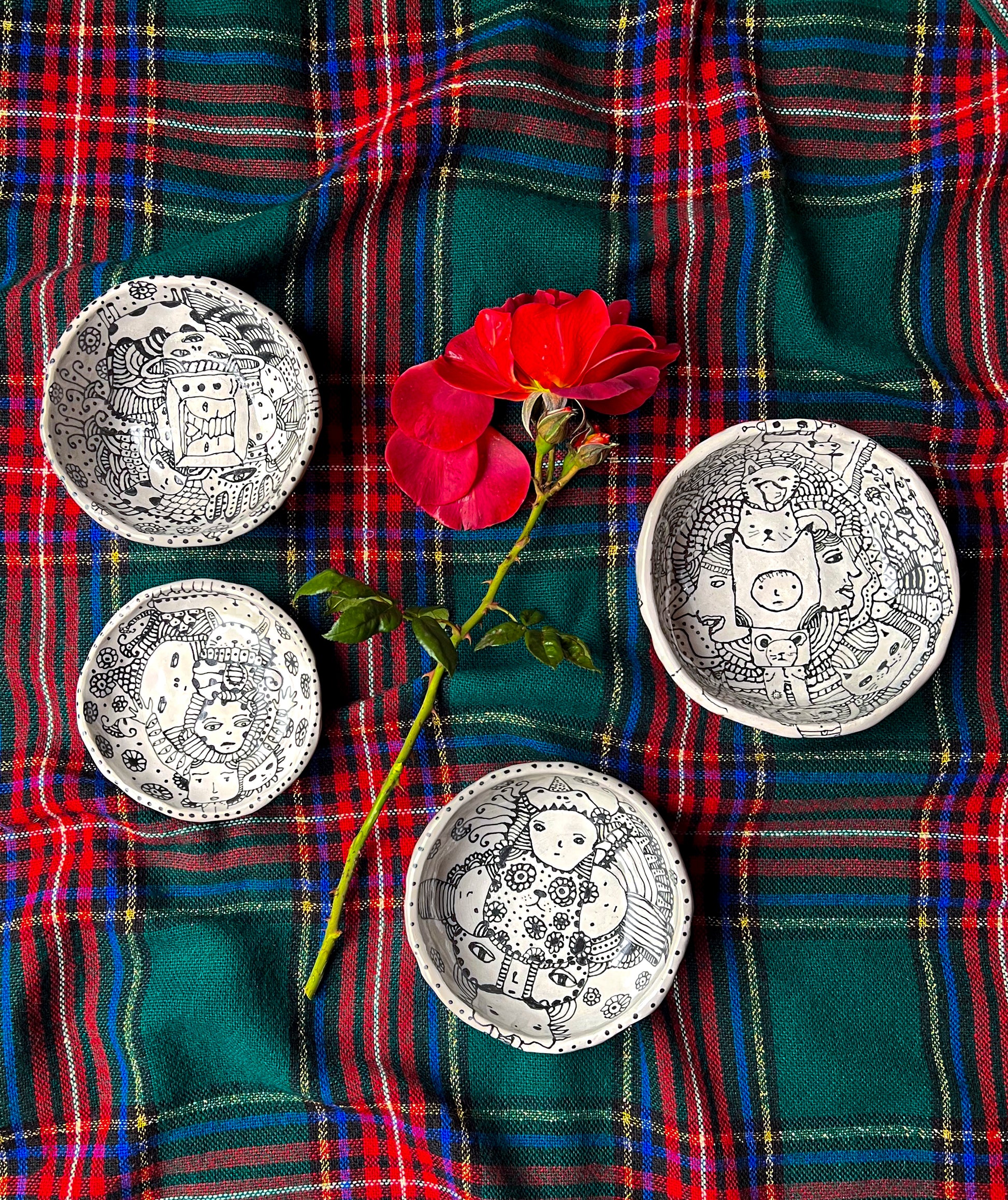 Four ceramic bowls shown together on plaid background with flower for scale with eclectic ink-drawn designs of animals, flowers, geometrics; artist Deb Blanco;  
