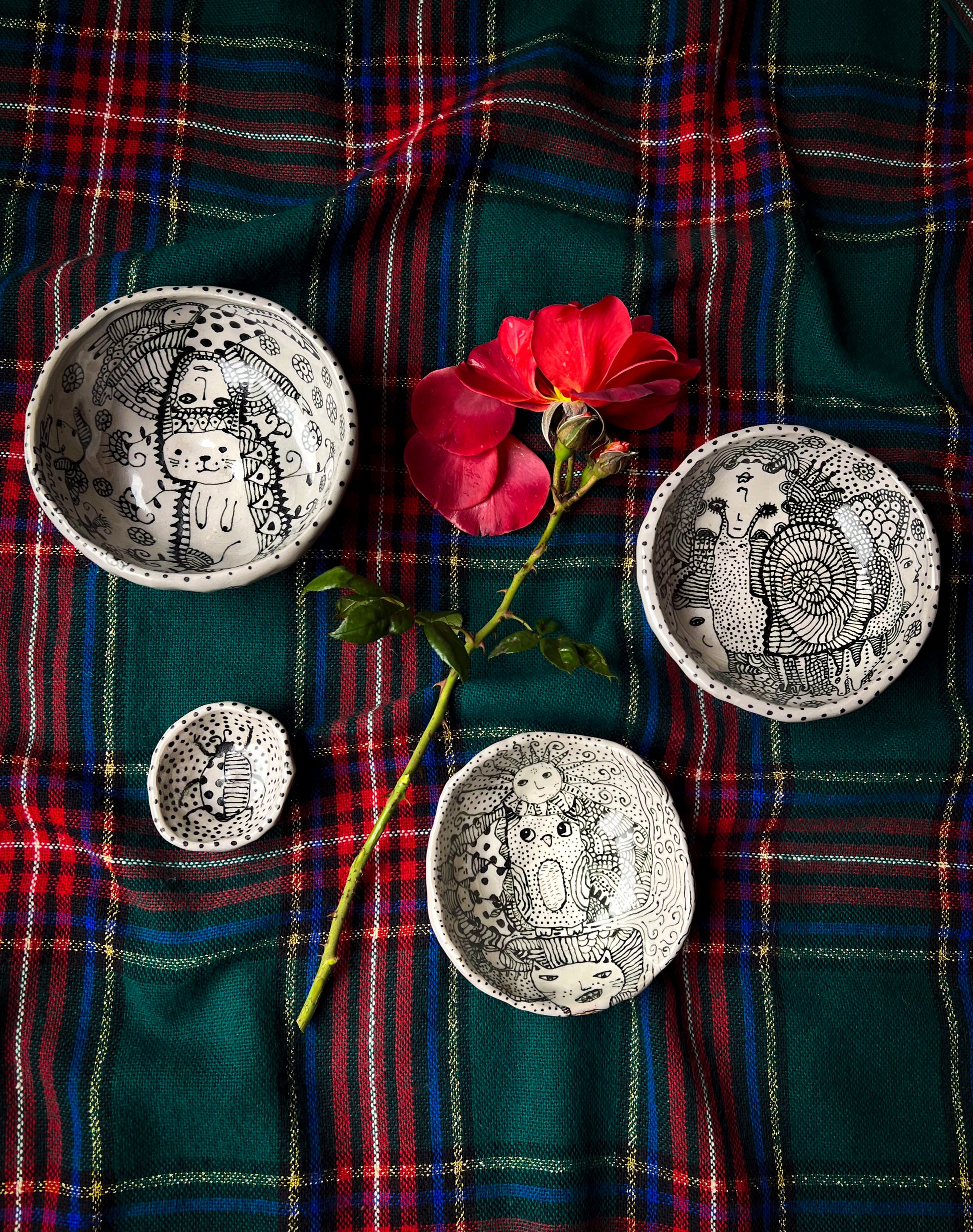 Four ceramic bowls shown together on plaid background with flower for scale with eclectic ink-drawn designs of animals, flowers, geometrics; artist Deb Blanco;  