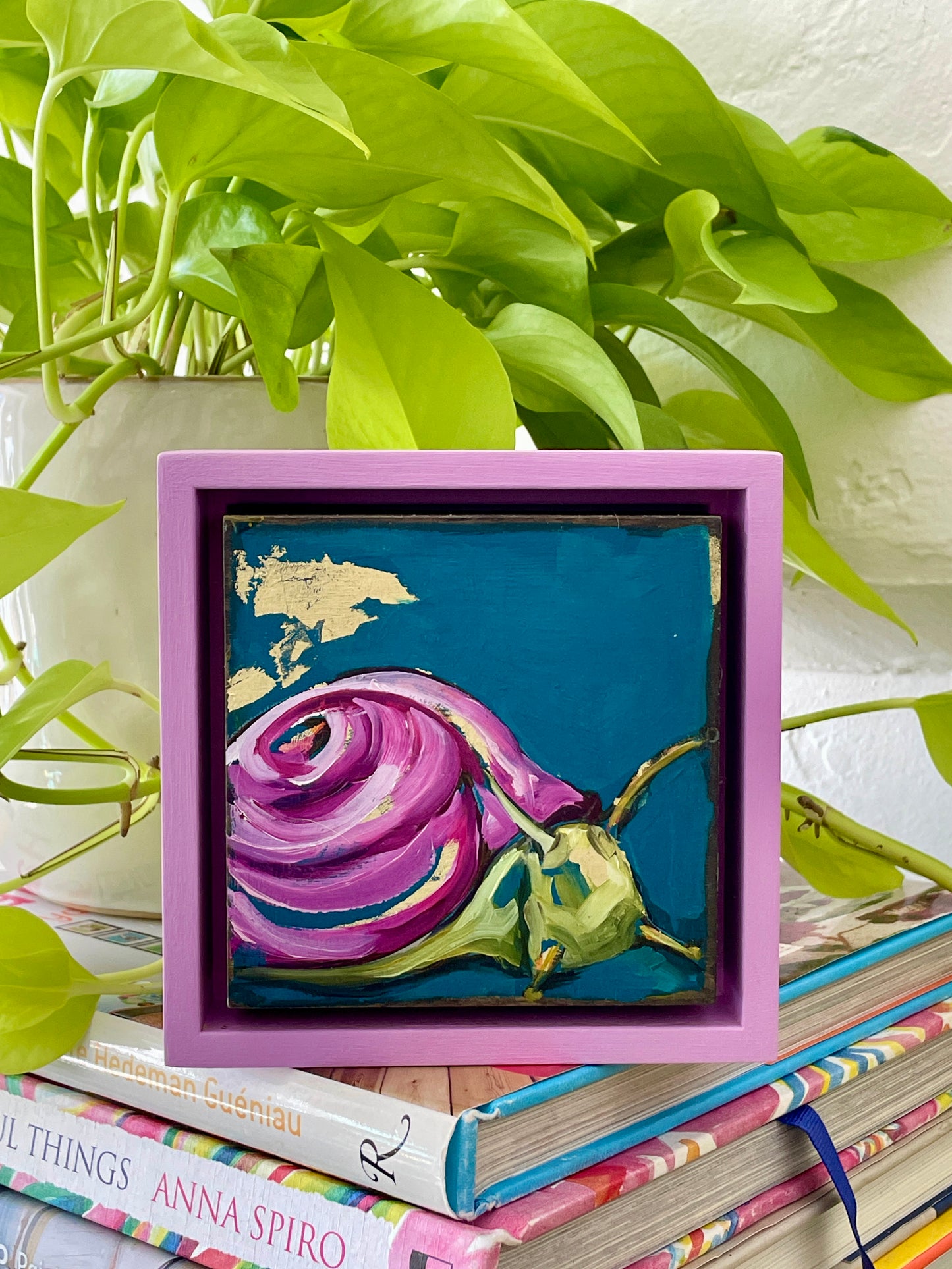 Colorful whimsical painting of a snail in blues and purples with touch of gold leaf; 5"x5" incl painted frame; artist Shaney Watters; shown sitting on stack of books