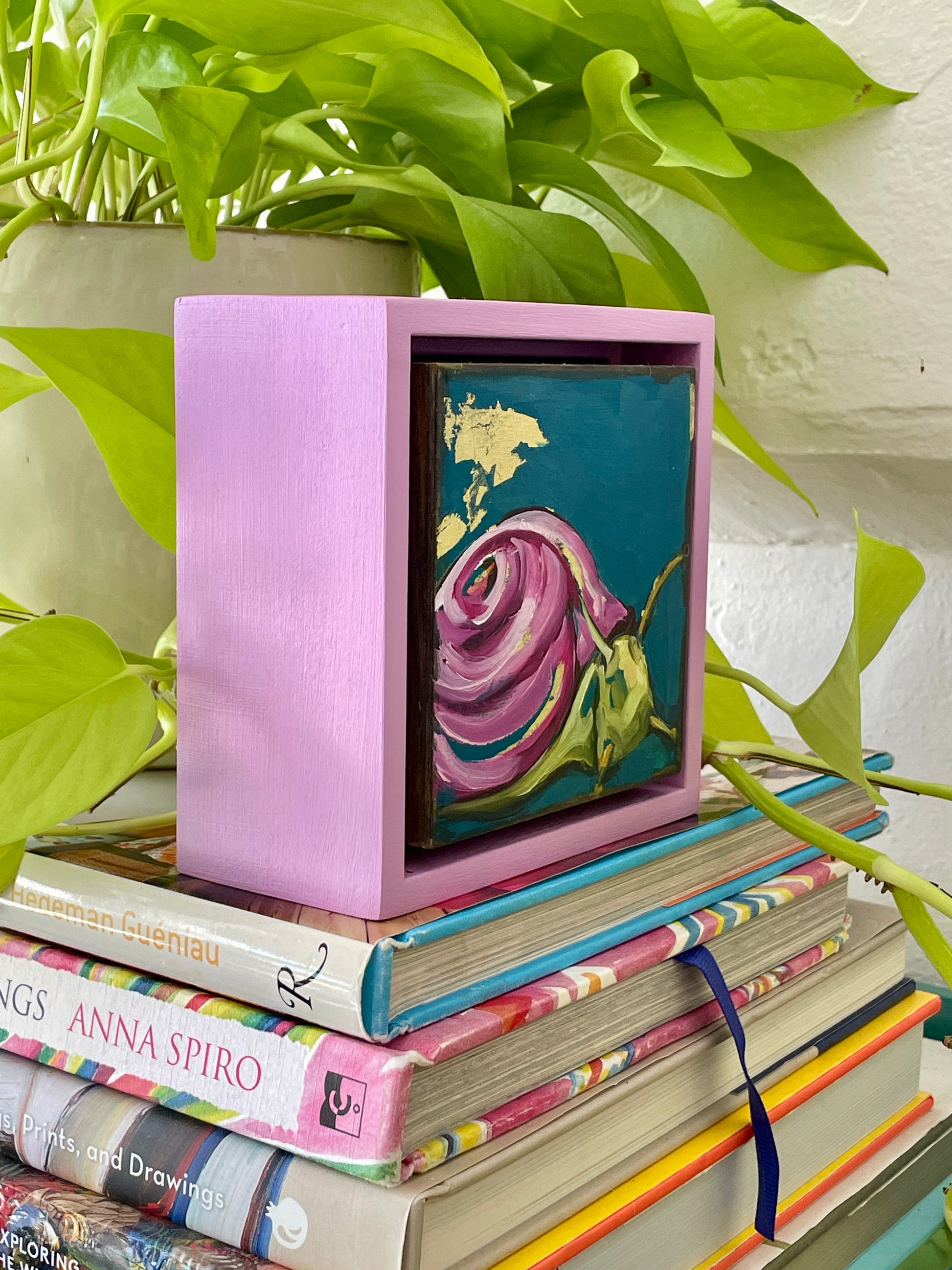 Colorful whimsical painting of a snail in blues and purples with touch of gold leaf; 5"x5" incl painted frame; artist Shaney Watters; shown side view displayed on stack of books