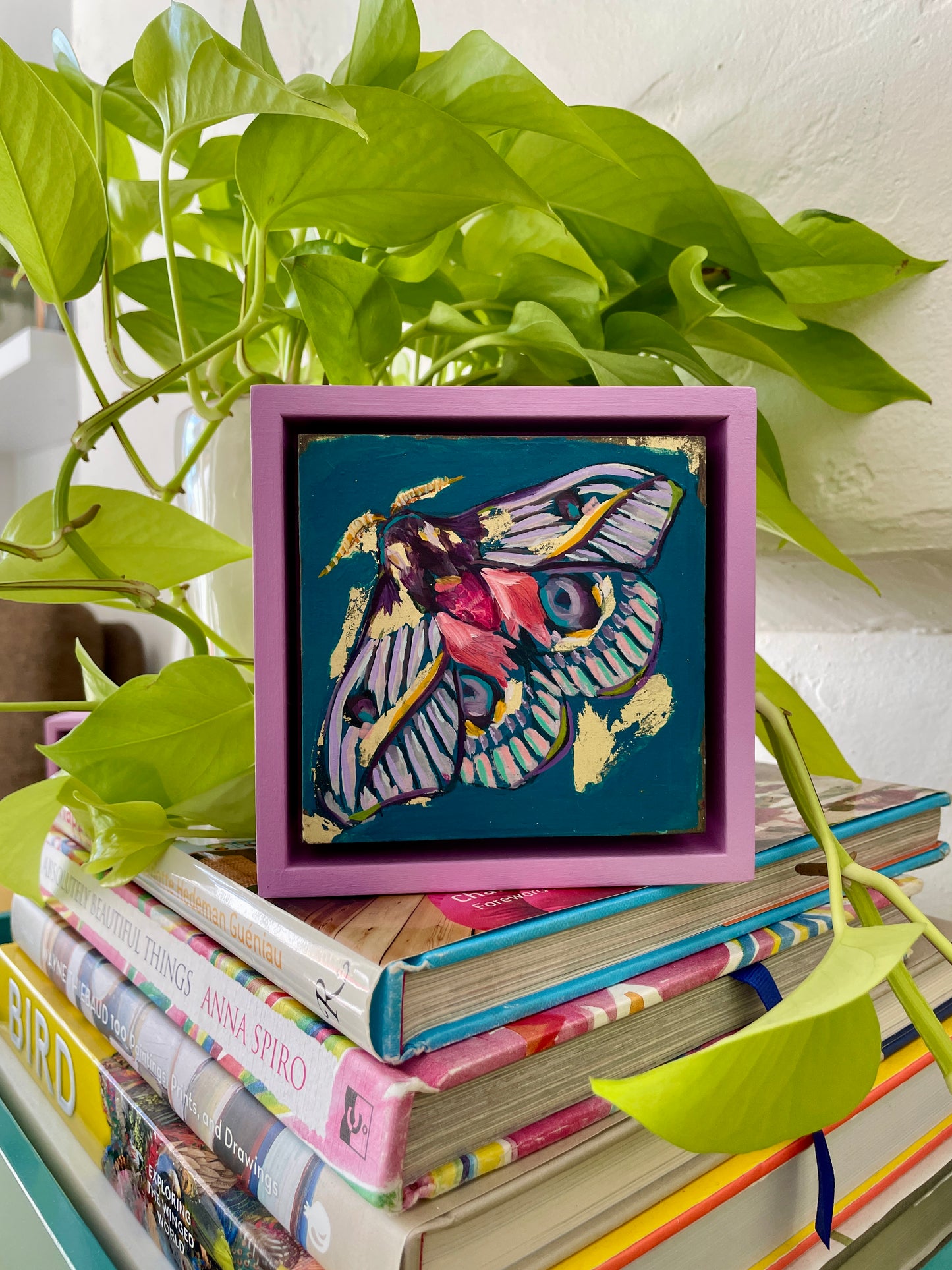 Colorful whimsical painting of a moth in blues, purples, pinks with touch of gold leaf; 5"x5" incl painted frame; artist Shaney Watters; shown sitting on top of stack of books