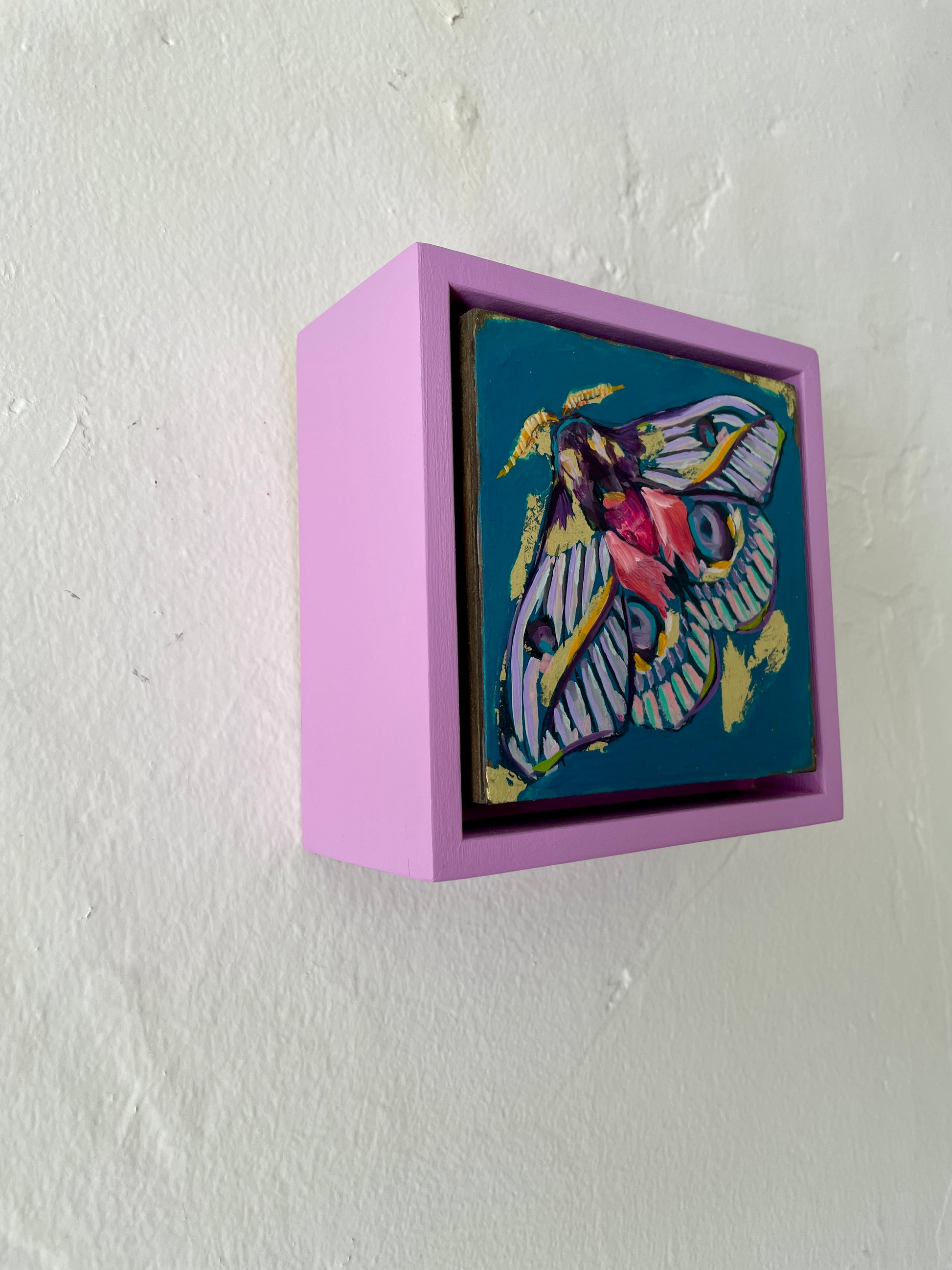 Colorful whimsical painting of a moth in blues, purples, pinks with touch of gold leaf; 5"x5" incl painted frame; artist Shaney Watters; side view shown hanging on wall