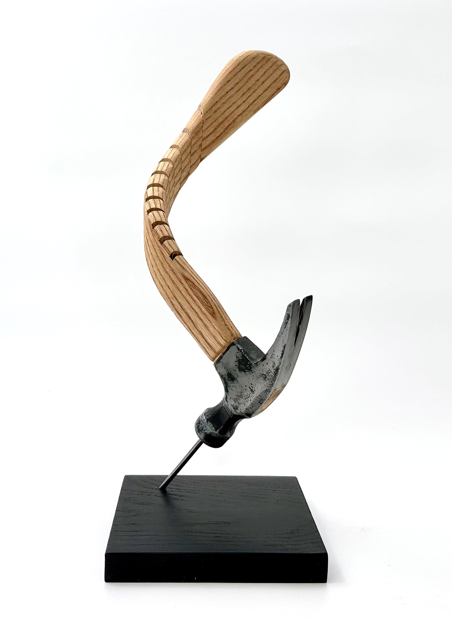 This exquisite sculpture is of a hammer resting at an angle on a nail that is attached the base. The hammer of the handle has been bend, using a series of small slits on its side. The slits allow the wood to curve and provide texture to the piece.