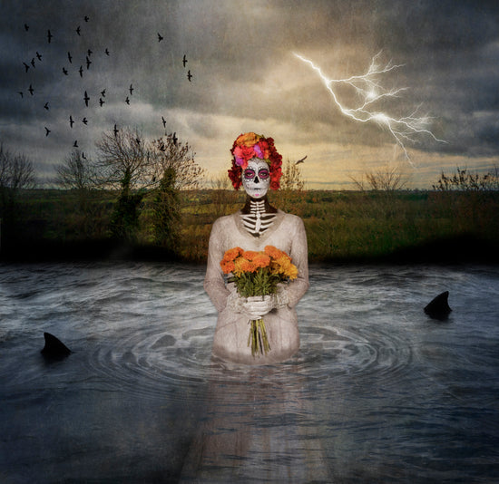 Digital composite image of a Catrina standing in water holding marigolds with sharks circling and lightning and birds in the sky;  Dia de Muertos; measure 10