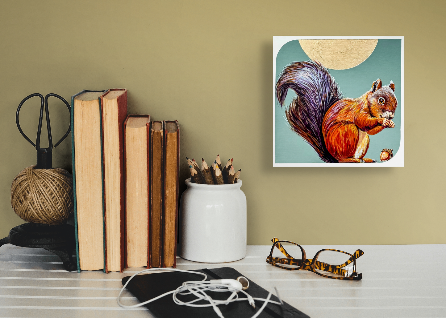 A Little Nutty hangs on a gold painted wall over a small table adorned with old books, a pencil holder and pair of glasses. 