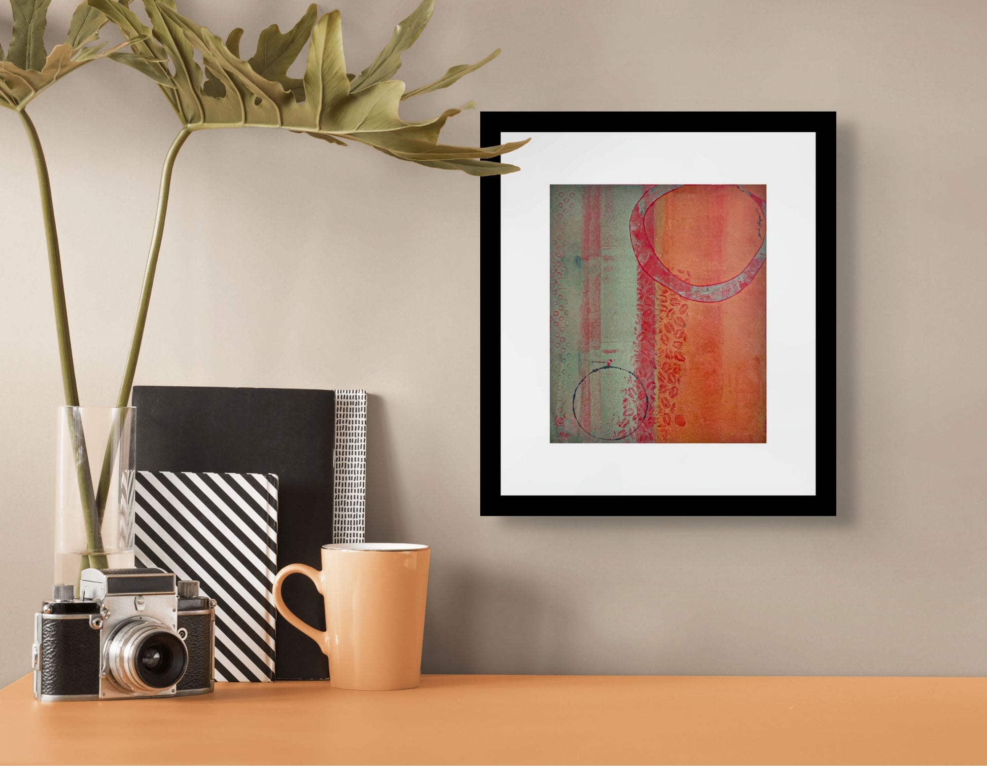 Abstract art piece with circular shapes and textured patterns using red/orange and green palette; frame w/white mat and black wood; artist Bob Hogue; shown in situ on beige wall over light brown table w/coffee cup, camera, several books and plant fronds in a vase