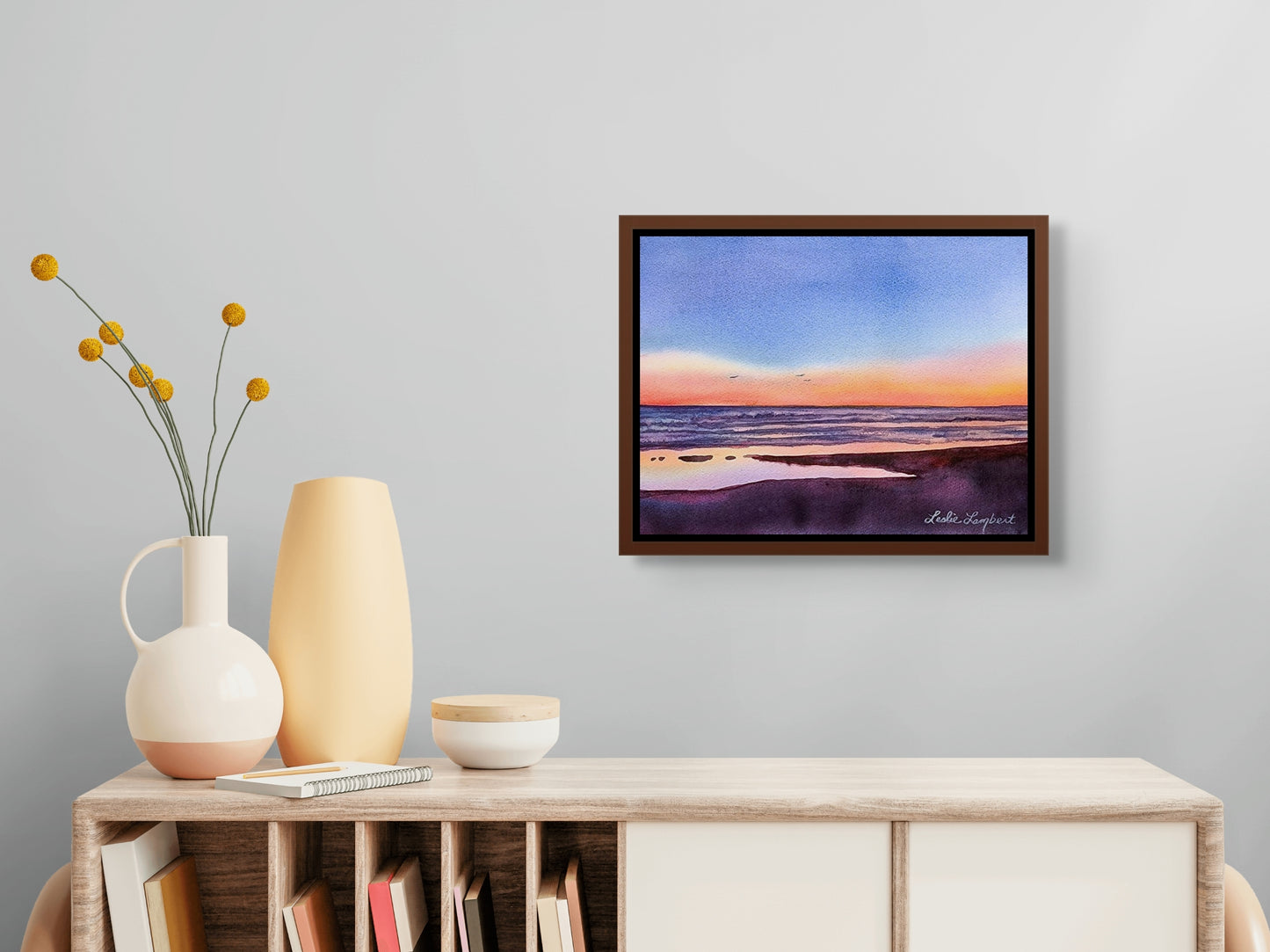 Beach Getaway is shown in a dark cherry wood frame situated over a light colored chest. On the chest sit a vase with flowers, an empty vase and a small bowl. 