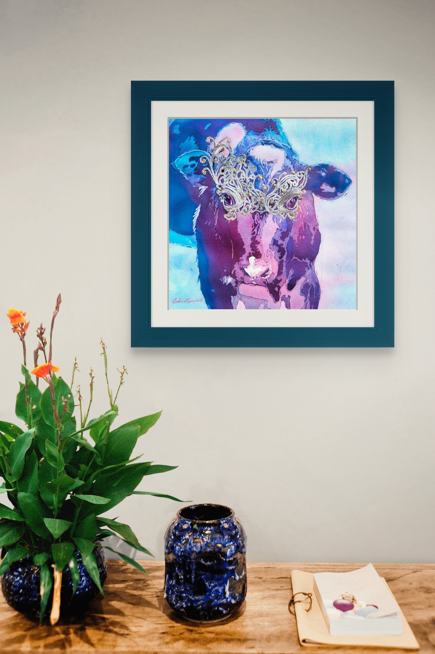 This in situ image shows Belle of the Ball hanging above a wooden table with a flower and vase. The painting, which is on cradle board, is shown in a blue green frame with a white mat. 